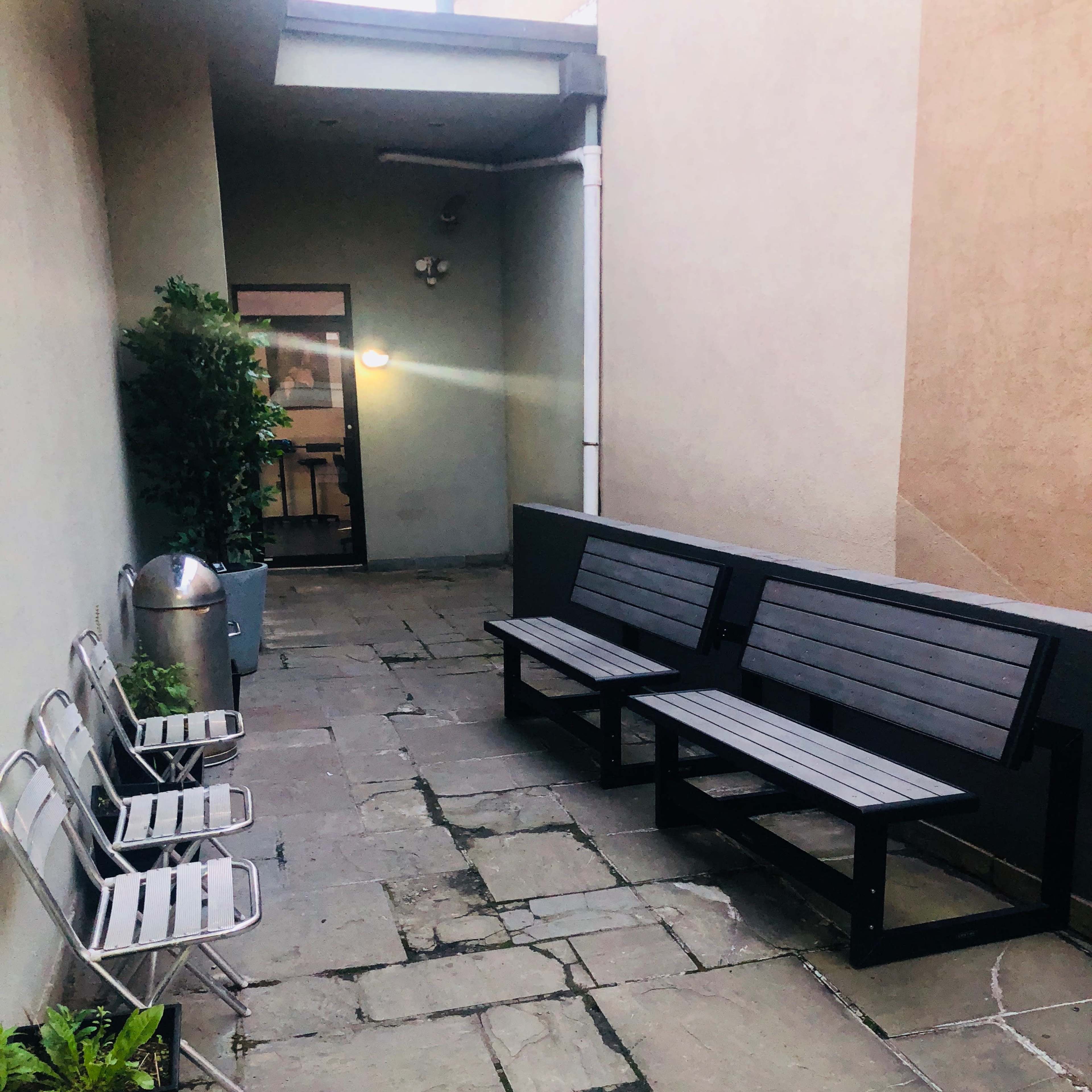 The scene shows a narrow corridor with stone flooring, two benches, a row of metal chairs, and planters along the walls.
