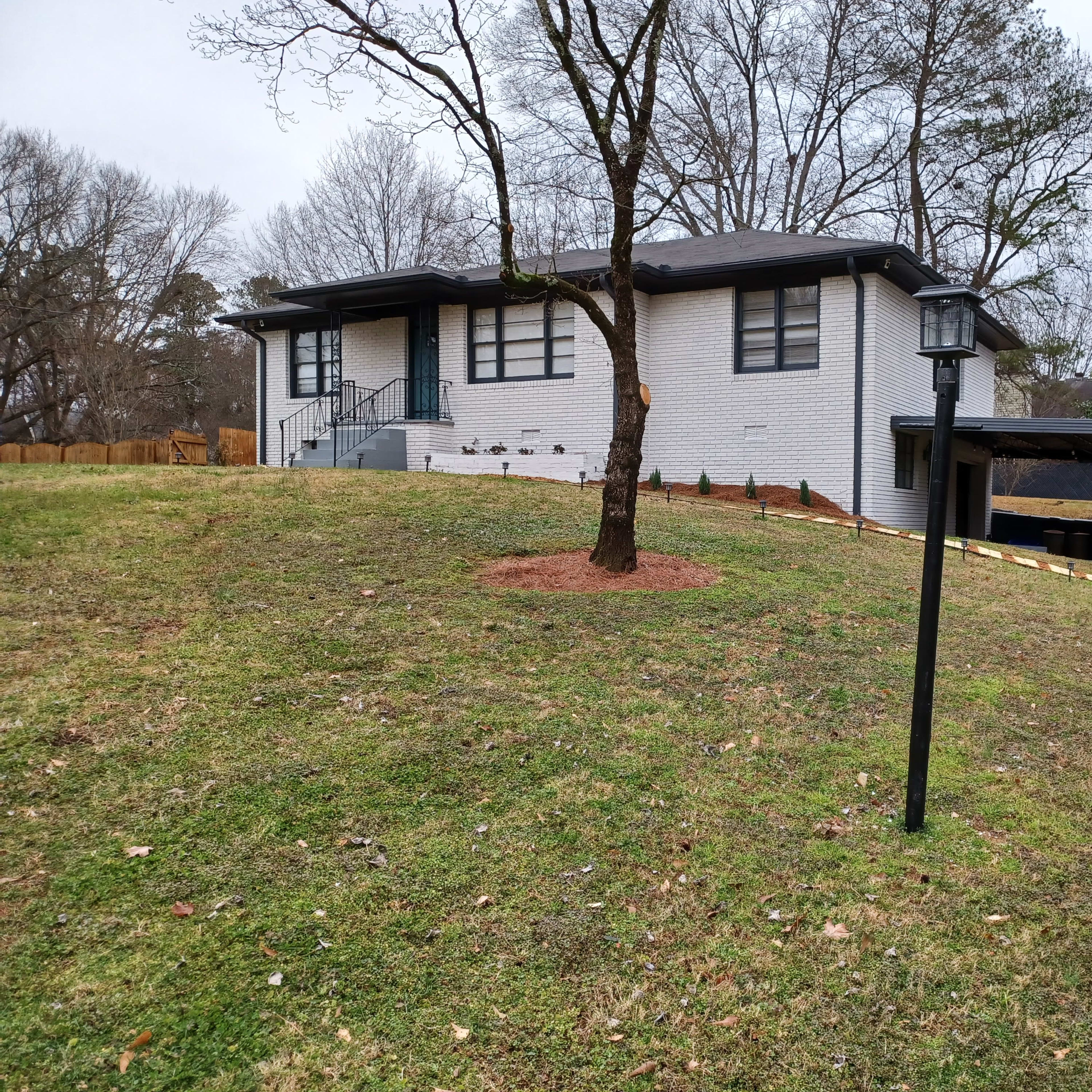 A single-story house with a white brick exterior, set on a grassy slope with a lamp post in the foreground and trees in the background.
