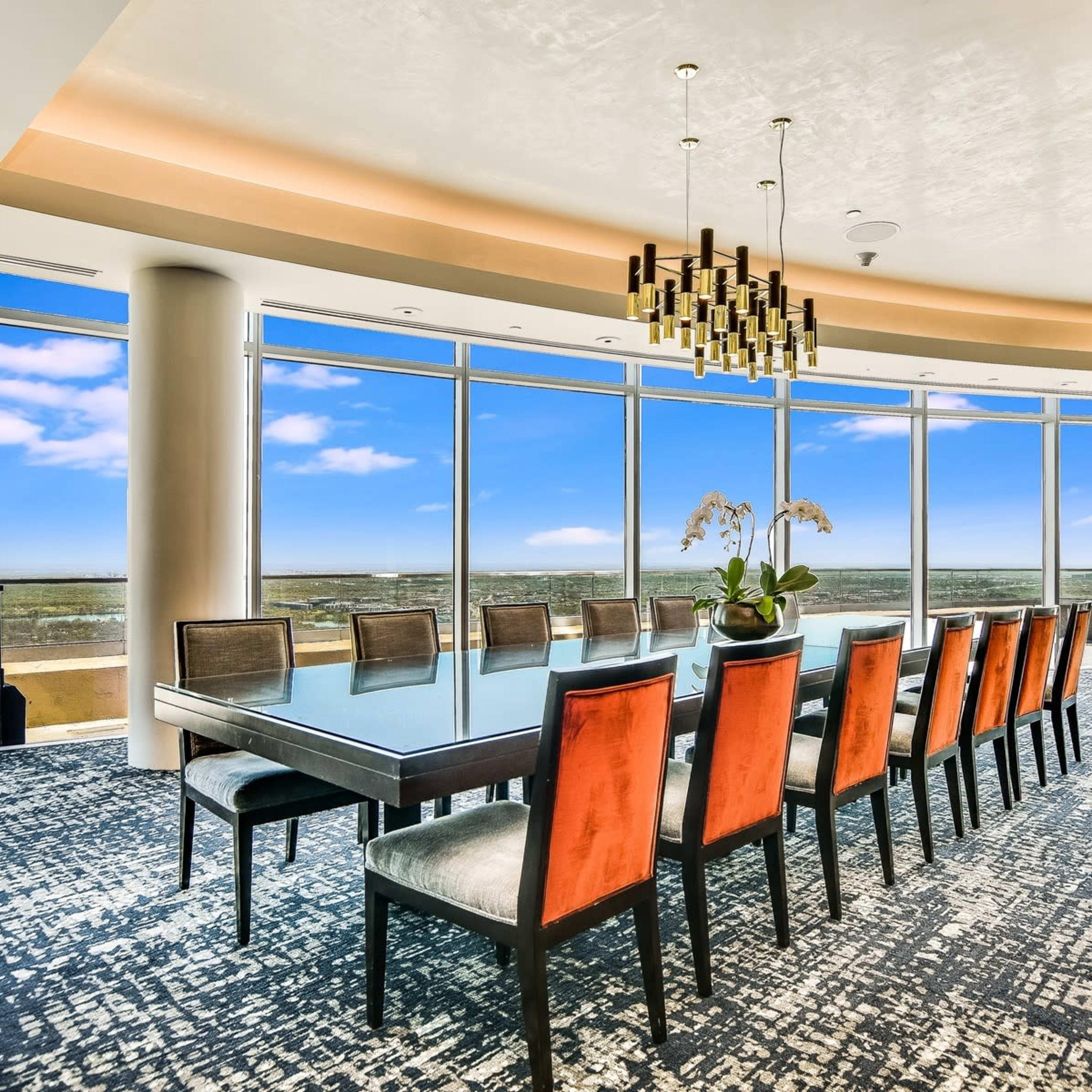 A large dining table is set with twelve chairs in a modern, brightly lit room featuring floor-to-ceiling windows that offer a view of the landscape outside.