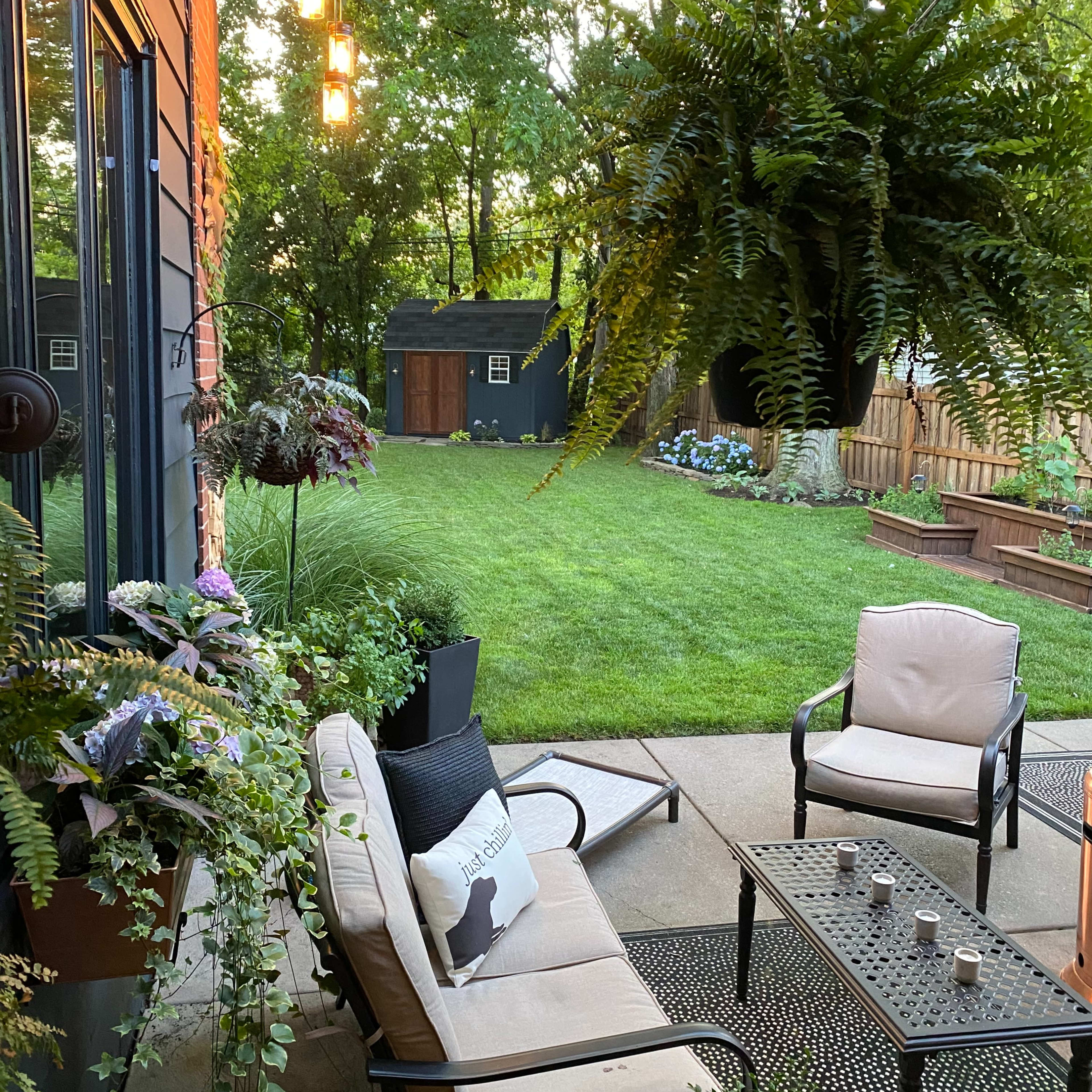 A cozy patio features two chairs and a table surrounded by hanging plants, leading to a green backyard with a shed and flower beds.