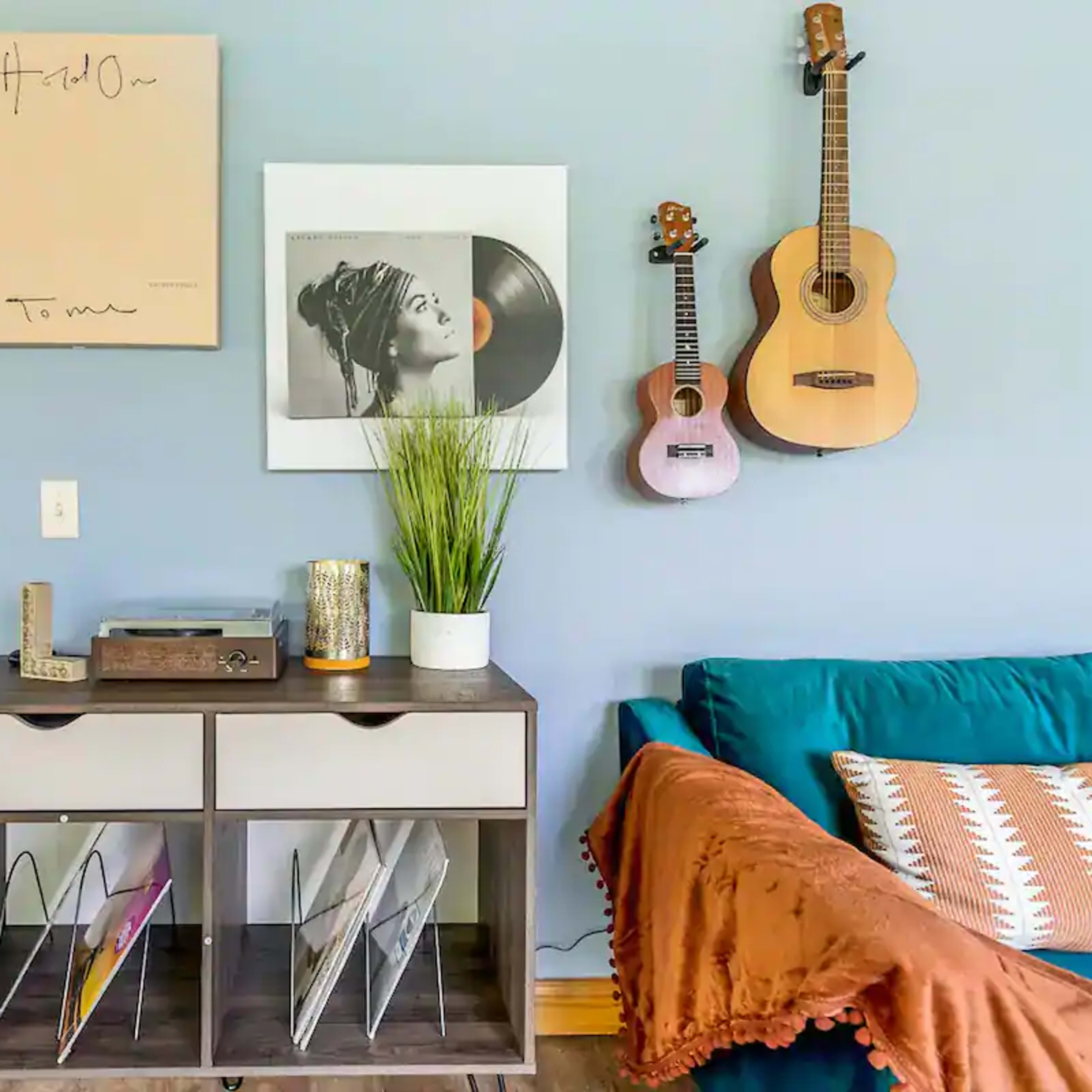 A console table with magazines, a record player, and decorative items is positioned against a light blue wall adorned with framed art and two guitars.