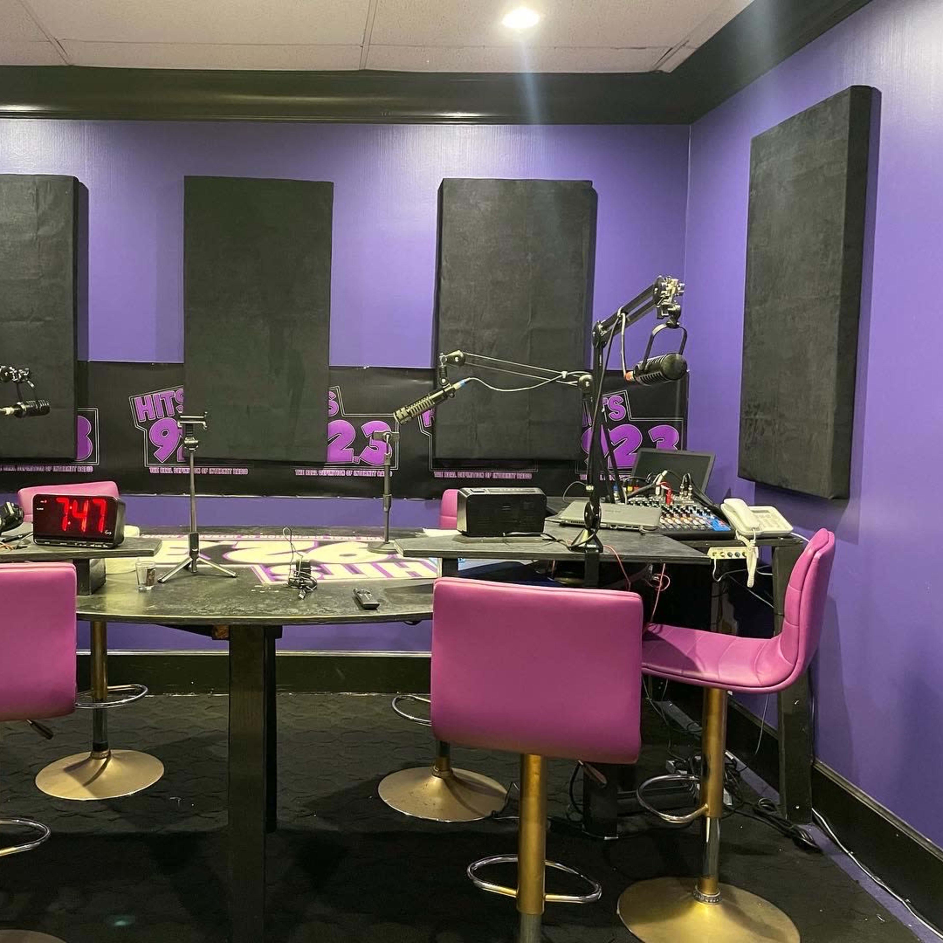 A radio studio with purple walls, two pink chairs, and equipment including microphones and a mixer on a central table.