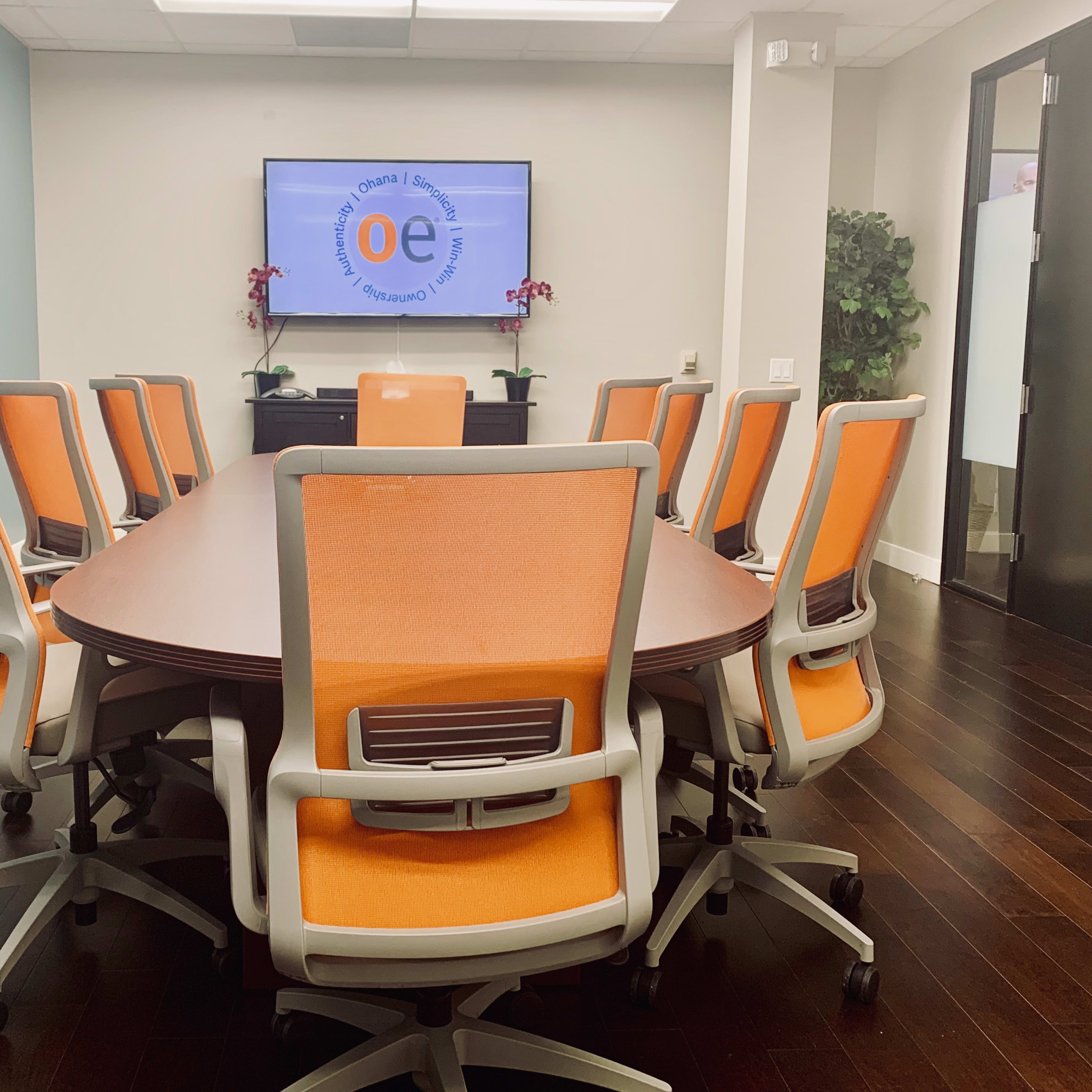 A conference room features a large oval table surrounded by ten orange and gray chairs, with a television displaying a logo on the wall.