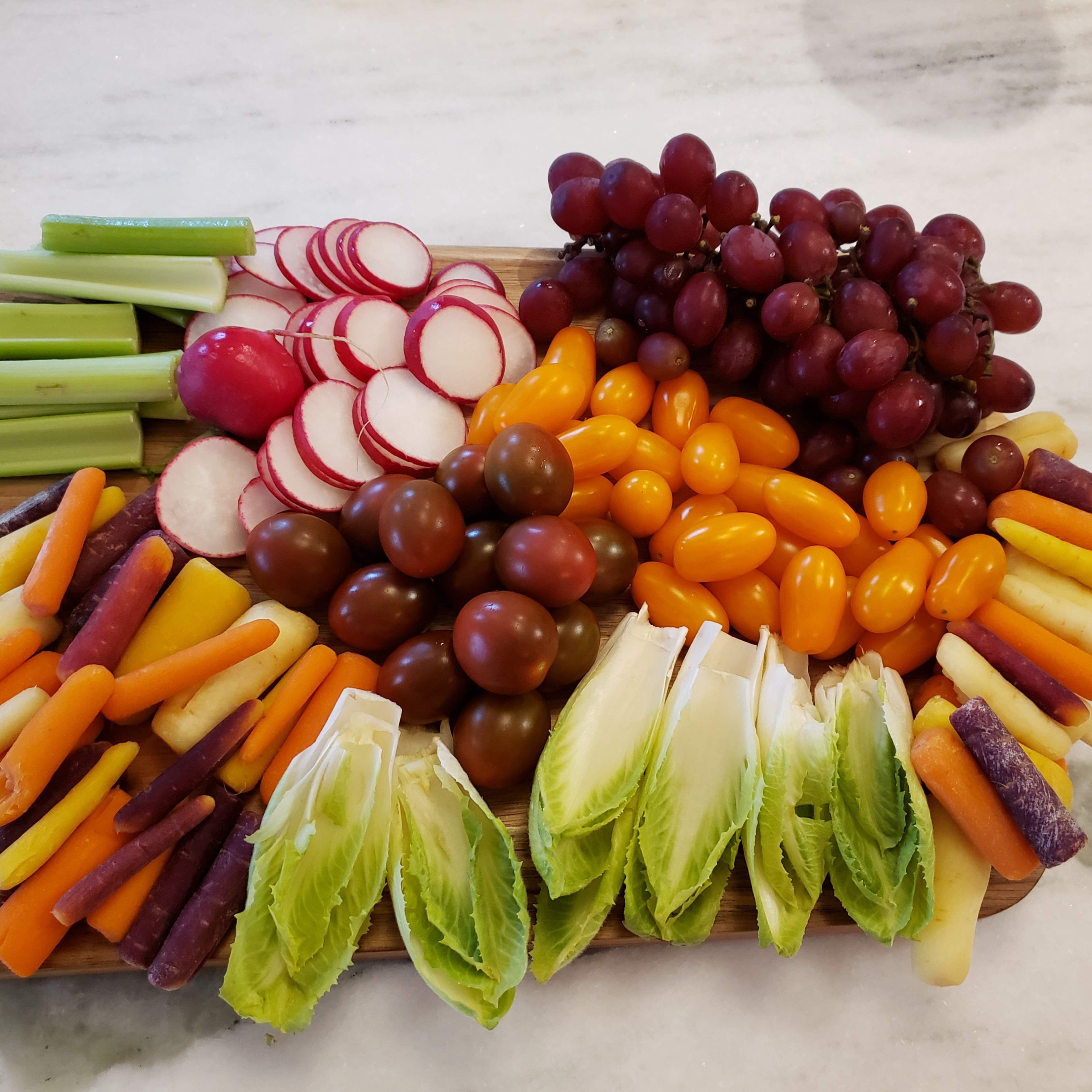 A wooden cutting board displays a colorful assortment of vegetables and grapes, including celery, radishes, grape tomatoes, grape varieties, and mixed carrots.