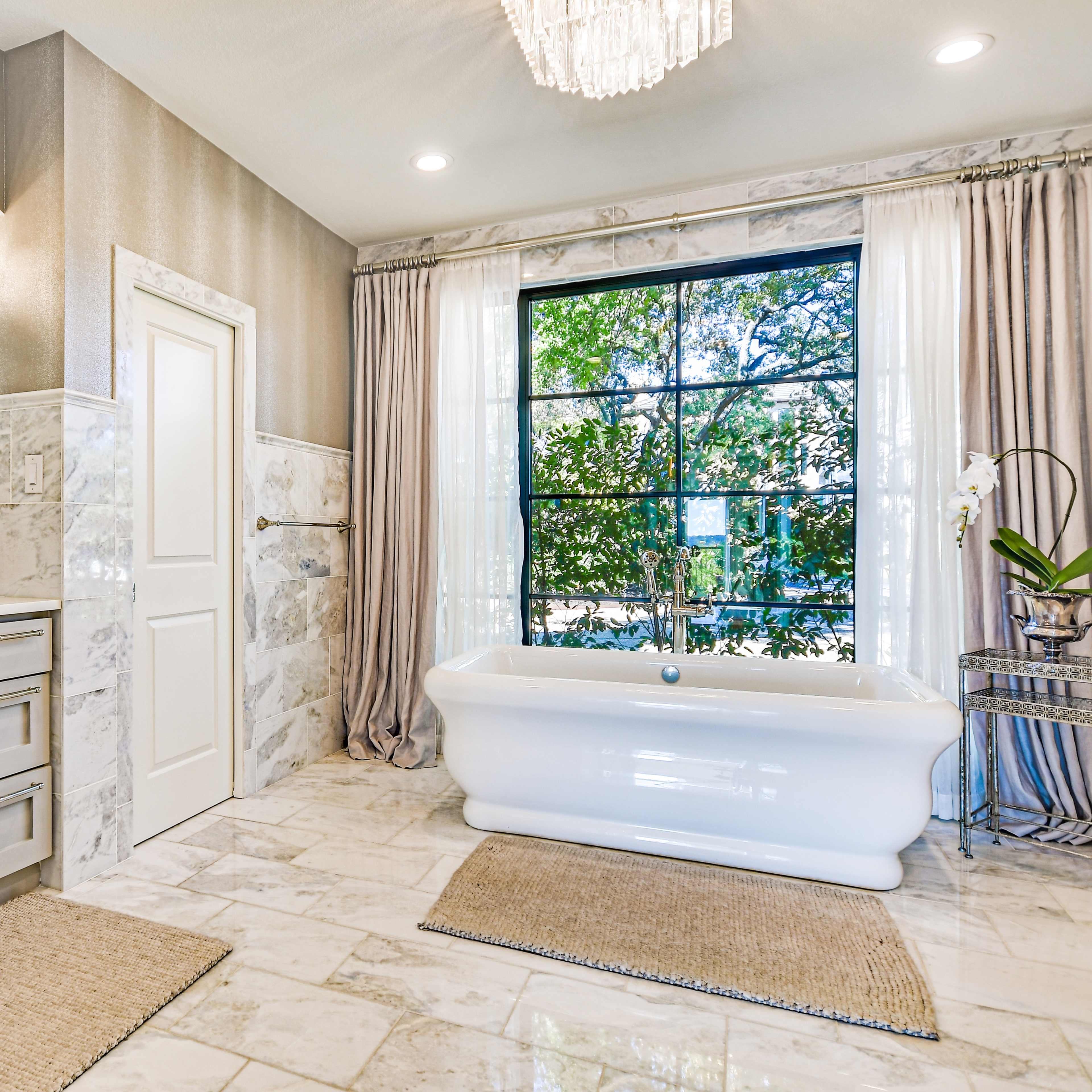 A modern bathroom features a freestanding bathtub, a double vanity, and large windows with a view of greenery.