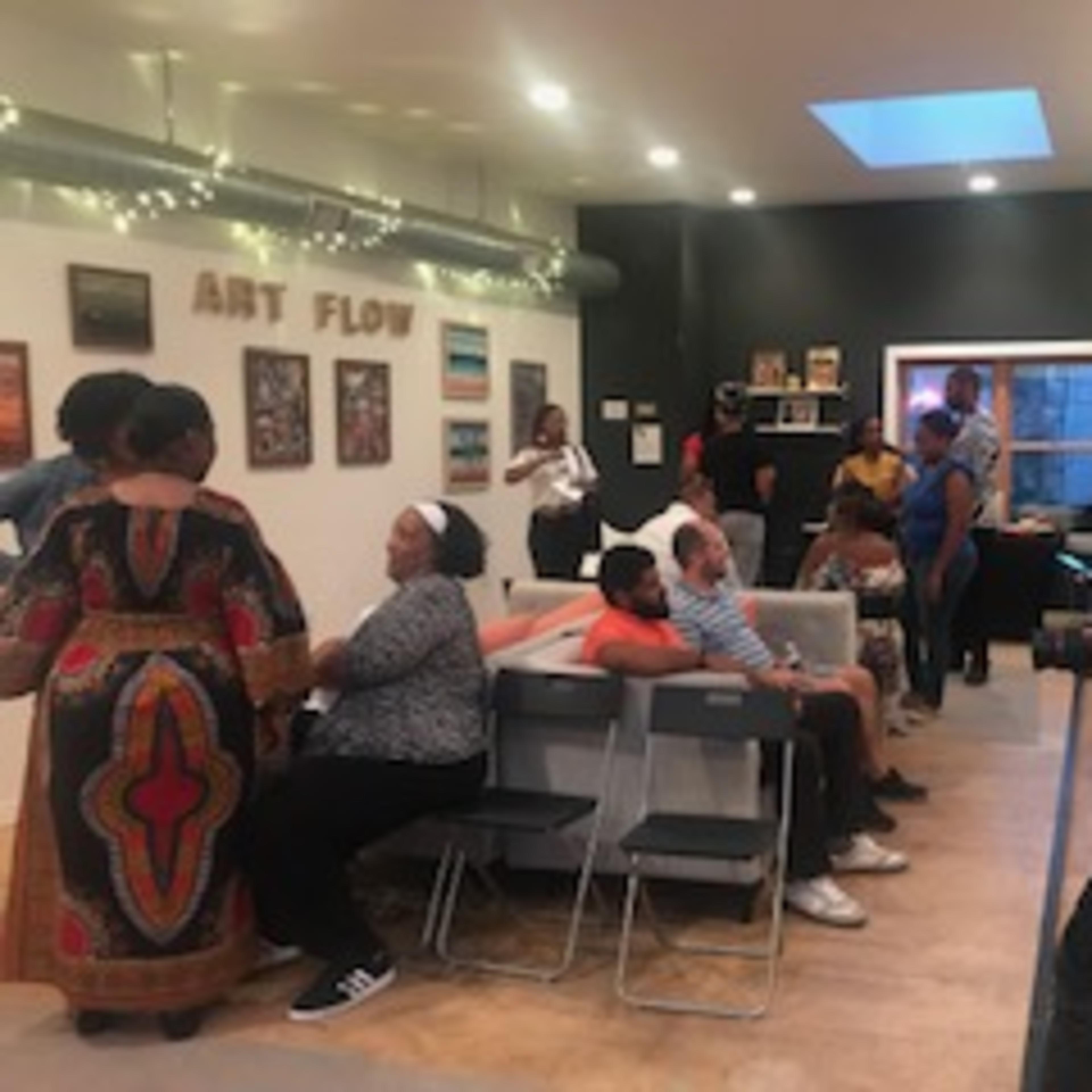 A group of people is gathered in an art space, engaging in conversation and taking photos, with artwork displayed on the walls.