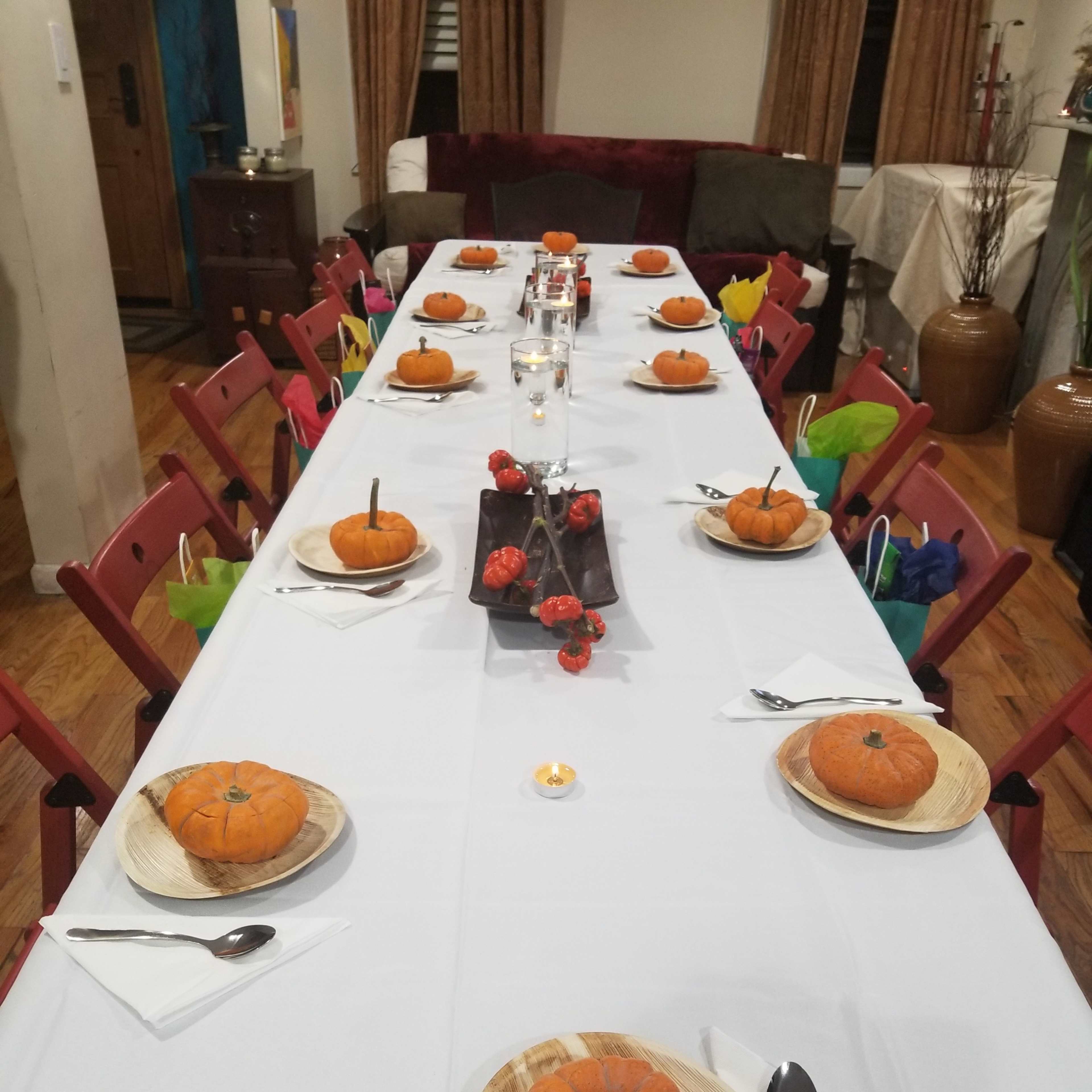 A long table is set with decorative pumpkins on wooden plates, surrounded by red folding chairs and a centerpiece of red decorations.