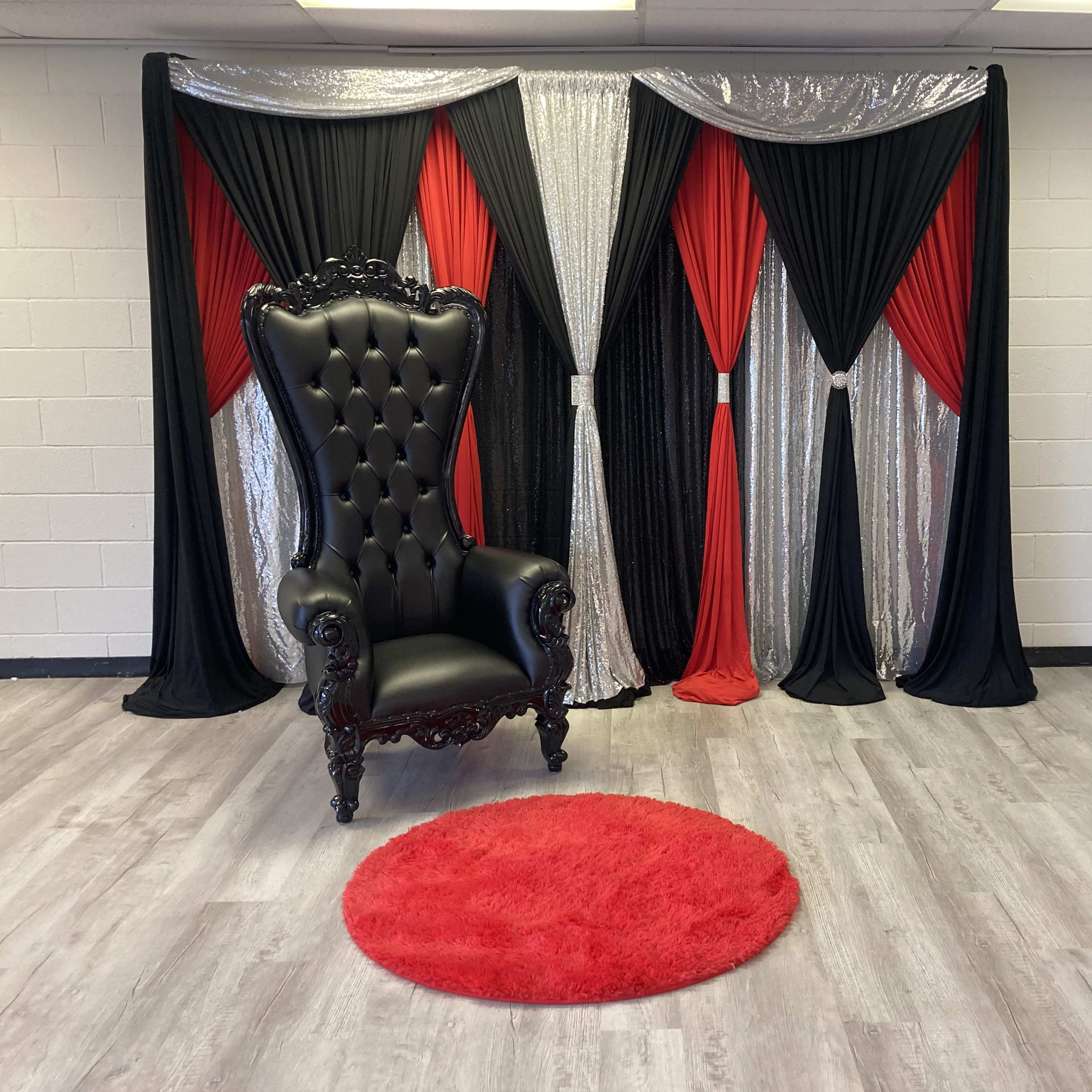 A black, tufted chair in front of a decorative backdrop of red, black, and silver drapes, with a round red rug on the floor.