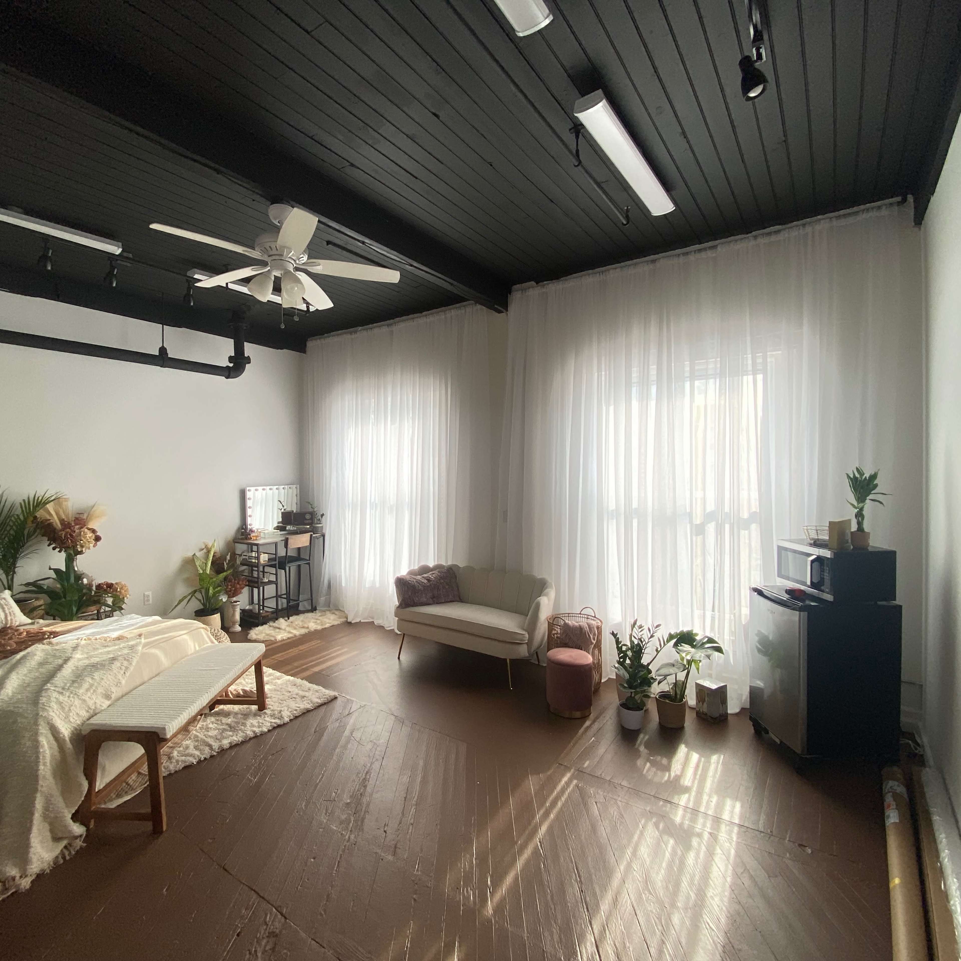 A spacious bedroom featuring a bed with light bedding, a couch, and several potted plants, illuminated by natural light through sheer curtains.