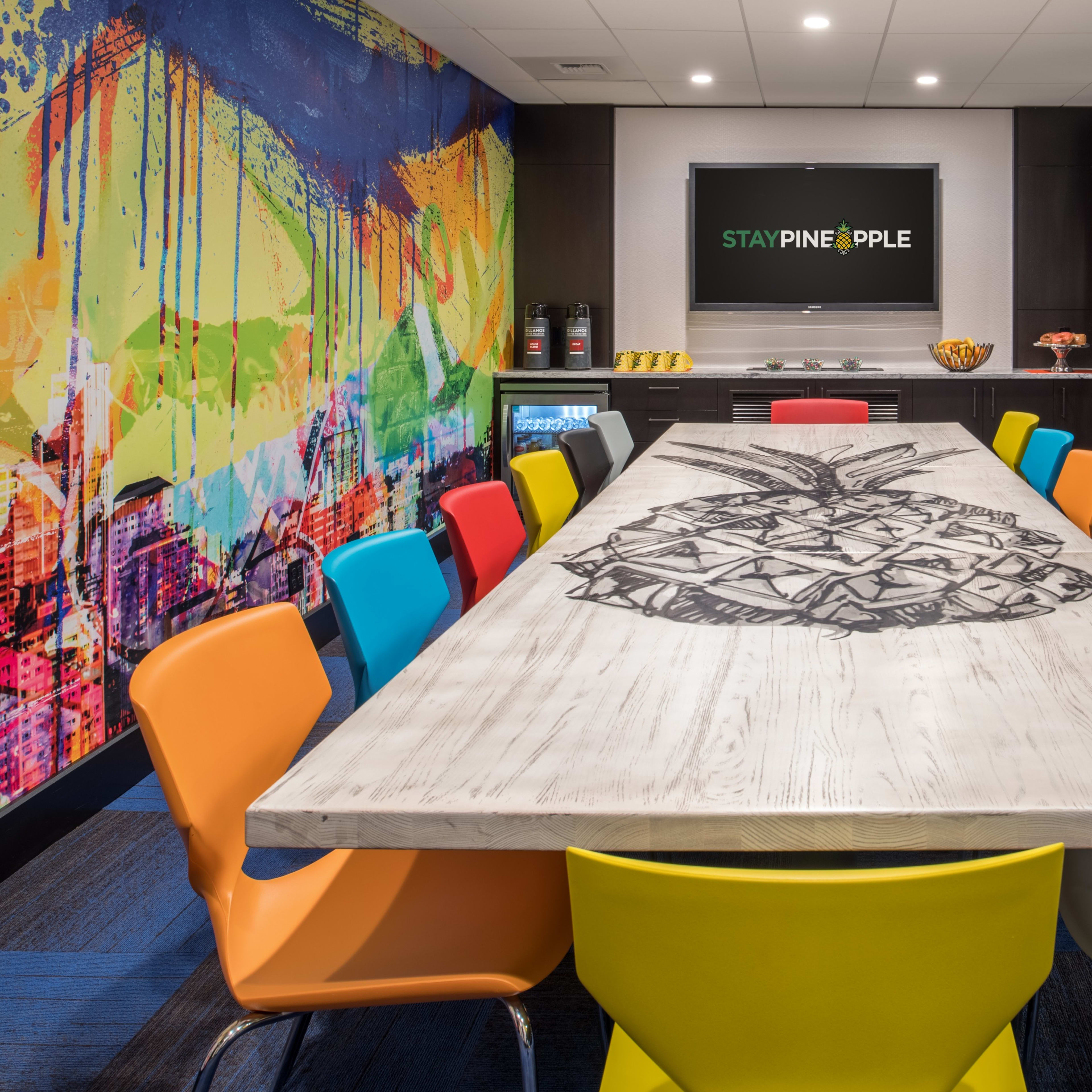 A conference room with a long table featuring a pineapple design, surrounded by colorful chairs and adorned with a vibrant wall mural.