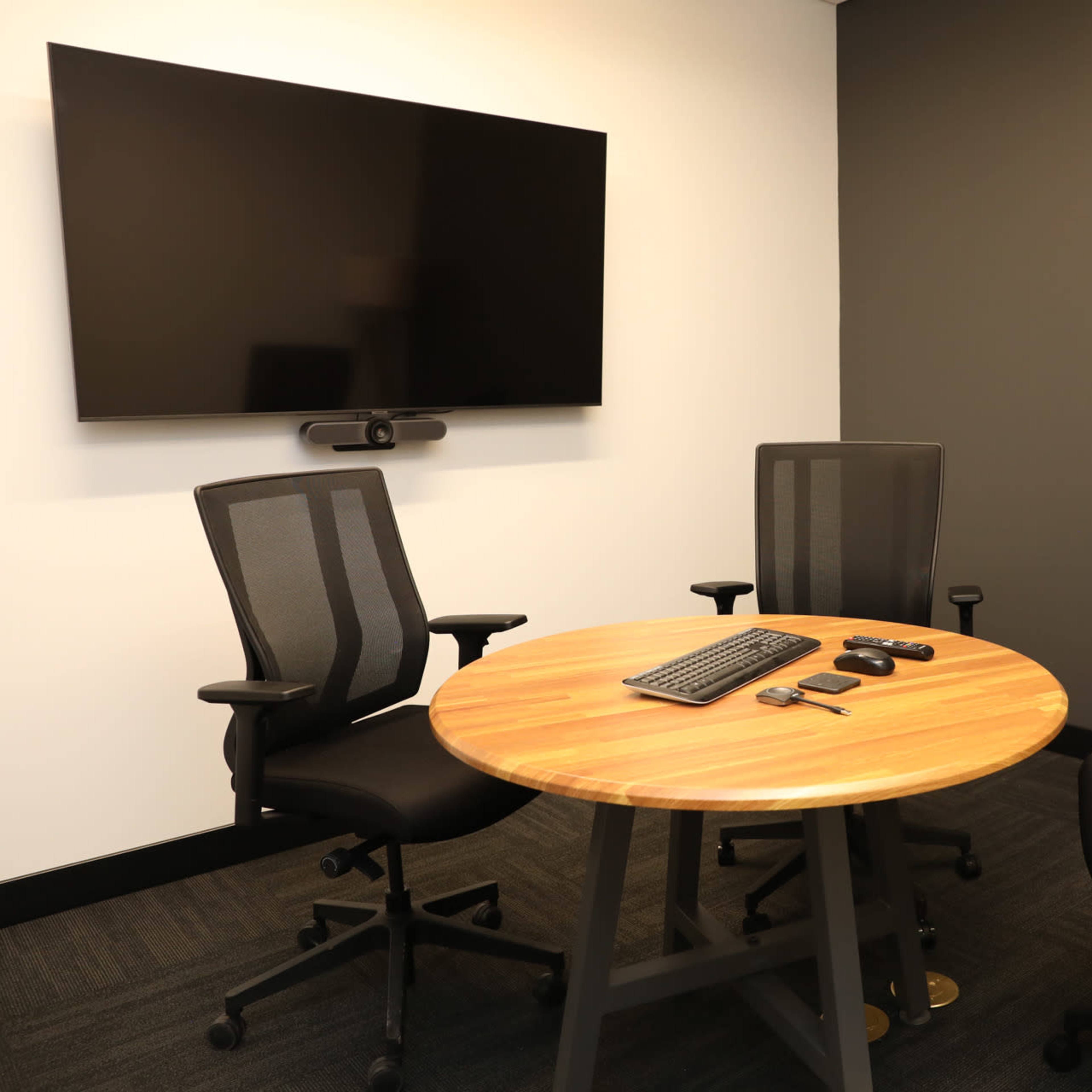 A small meeting room features a circular wooden table with three black chairs and a large wall-mounted screen.