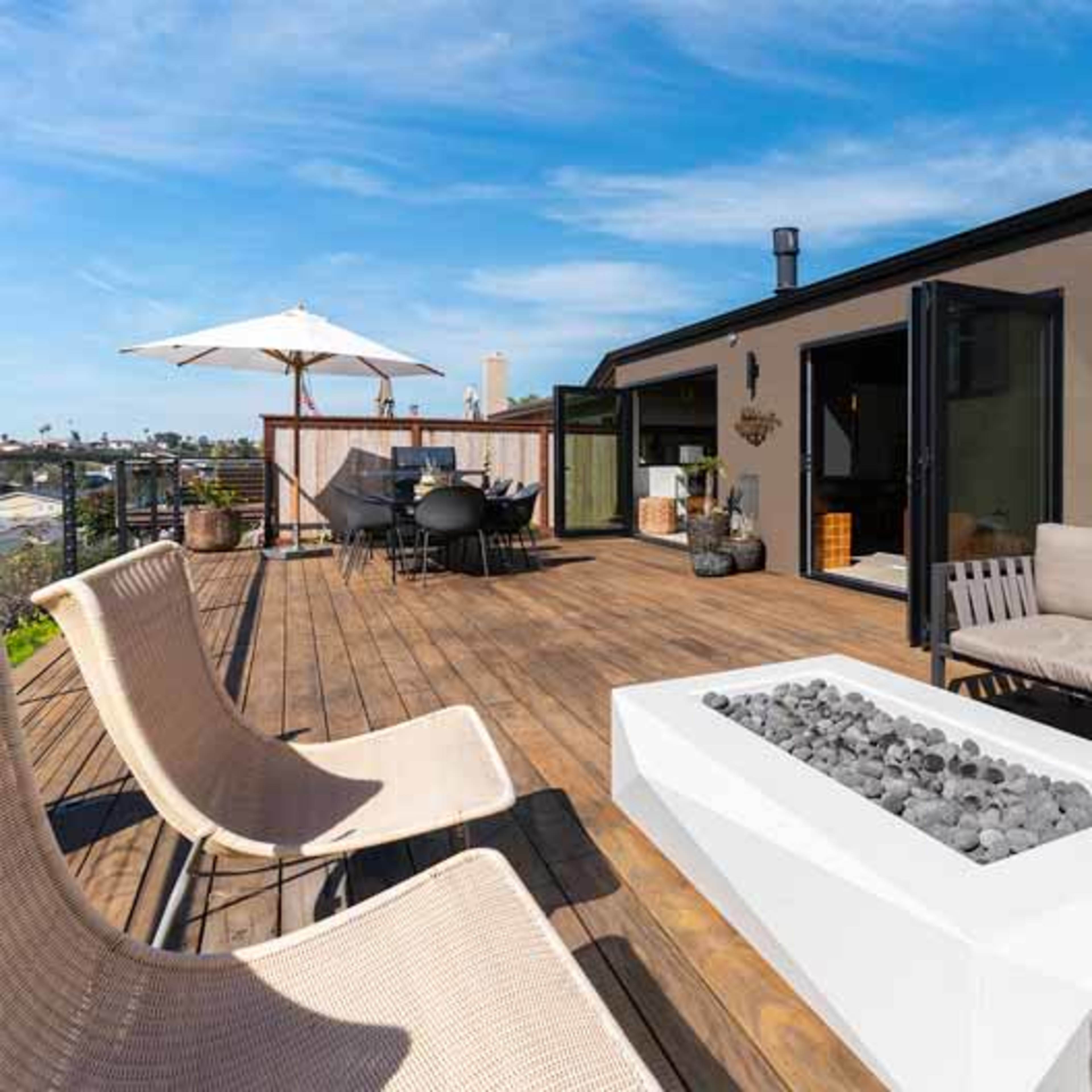 A modern rooftop terrace features a white fire pit surrounded by comfortable seating, with a dining area and an umbrella in the background.