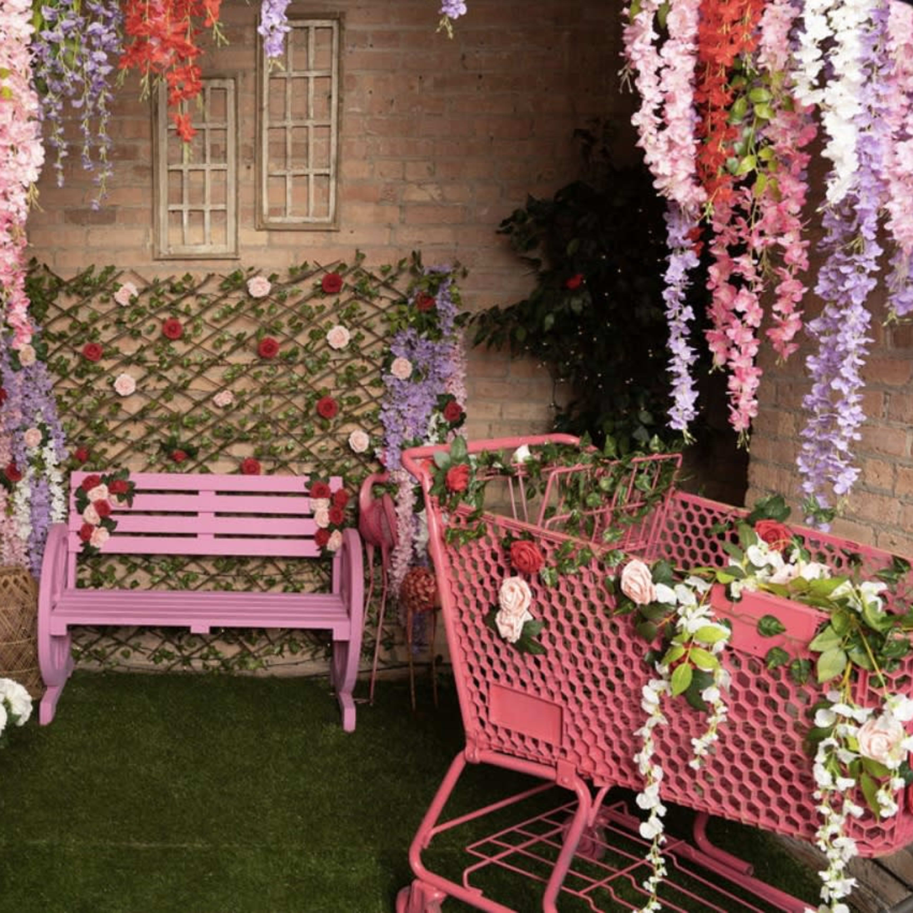 The scene features a decorated corner with a pink bench, two pink shopping carts adorned with flowers, and vibrant hanging floral arrangements against a brick wall.