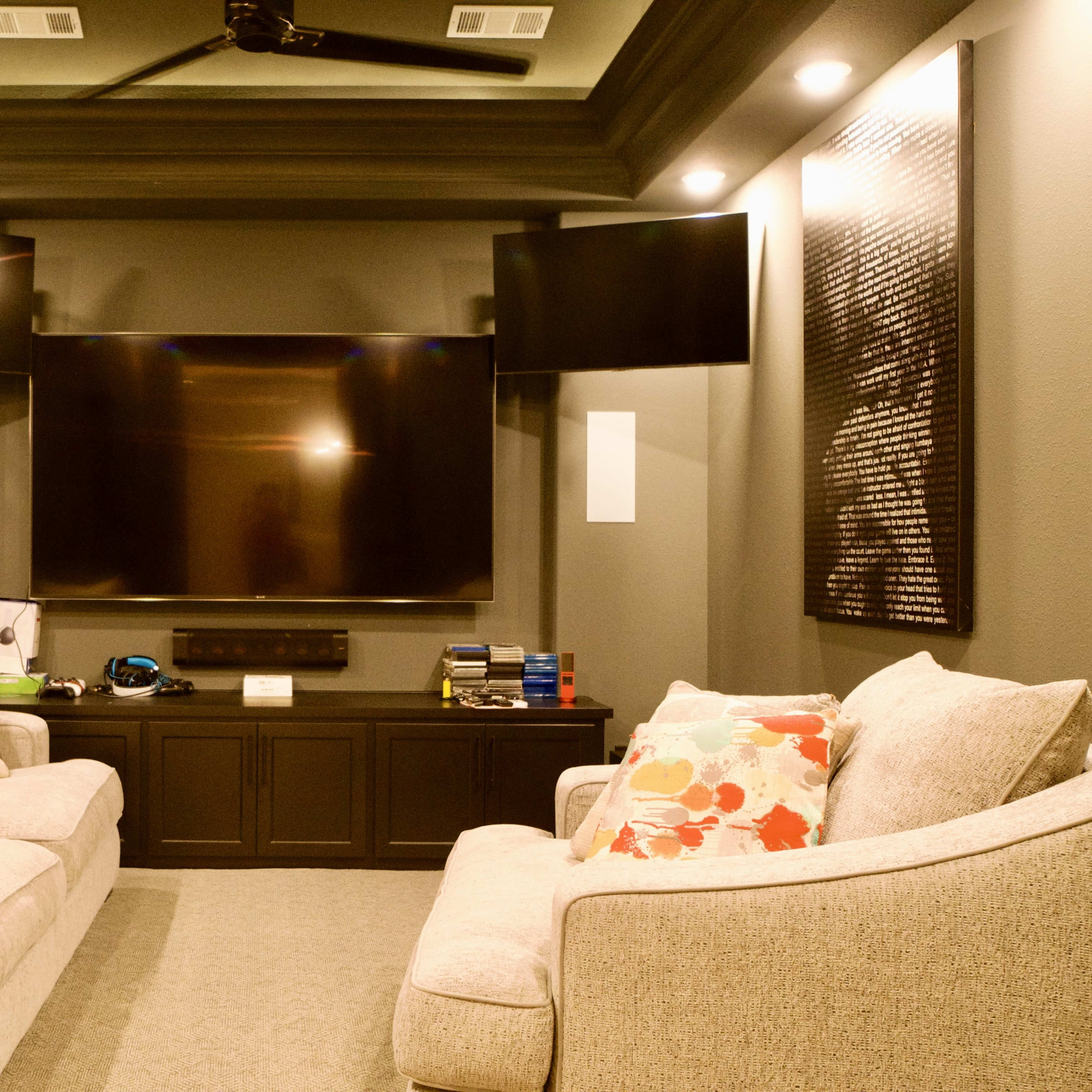 A small home theater room features a large flat-screen TV mounted on the wall, flanked by two smaller screens, with two couches facing the television.
