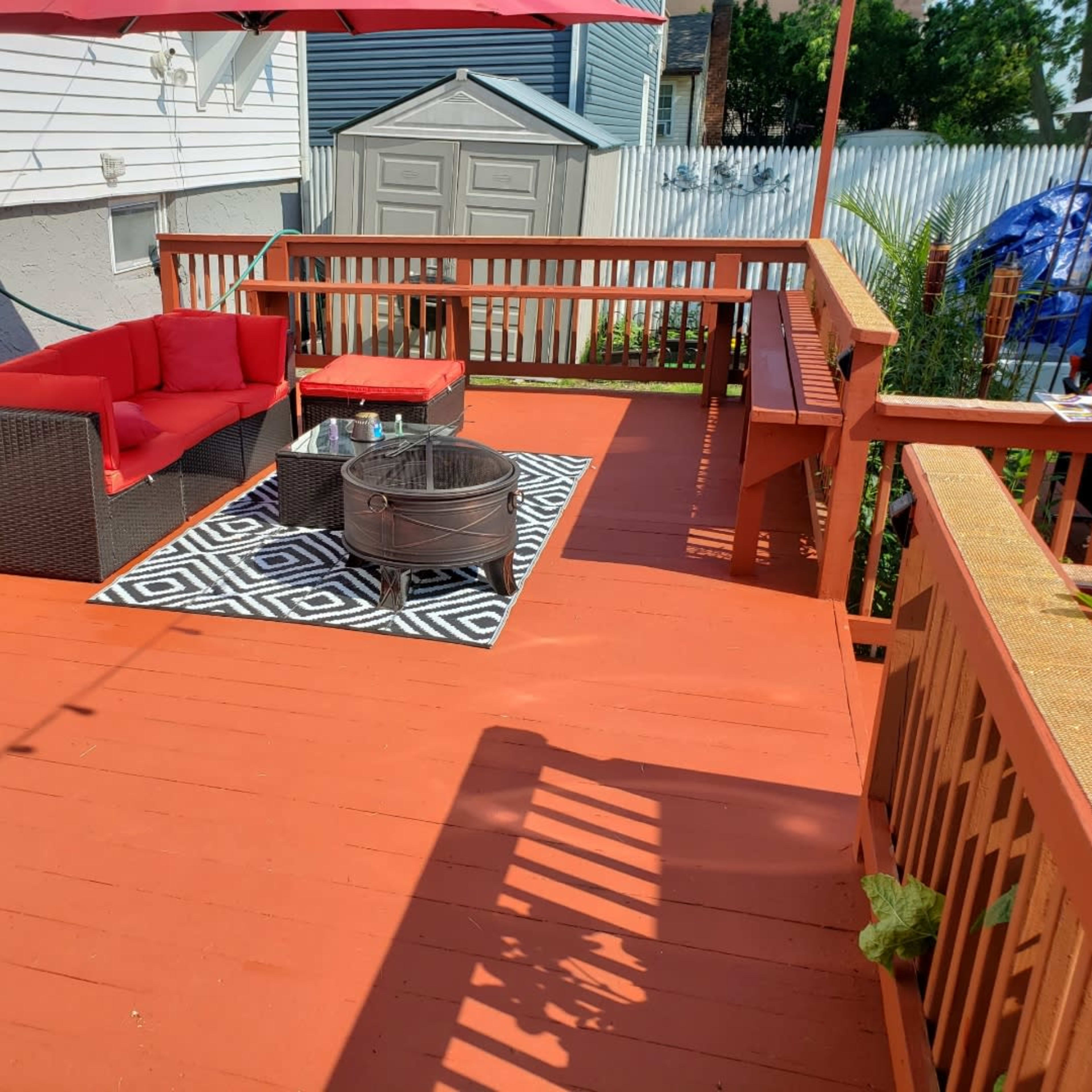 A red-painted wooden deck with a seating area featuring a red sofa and a circular fire pit, surrounded by a patterned rug.