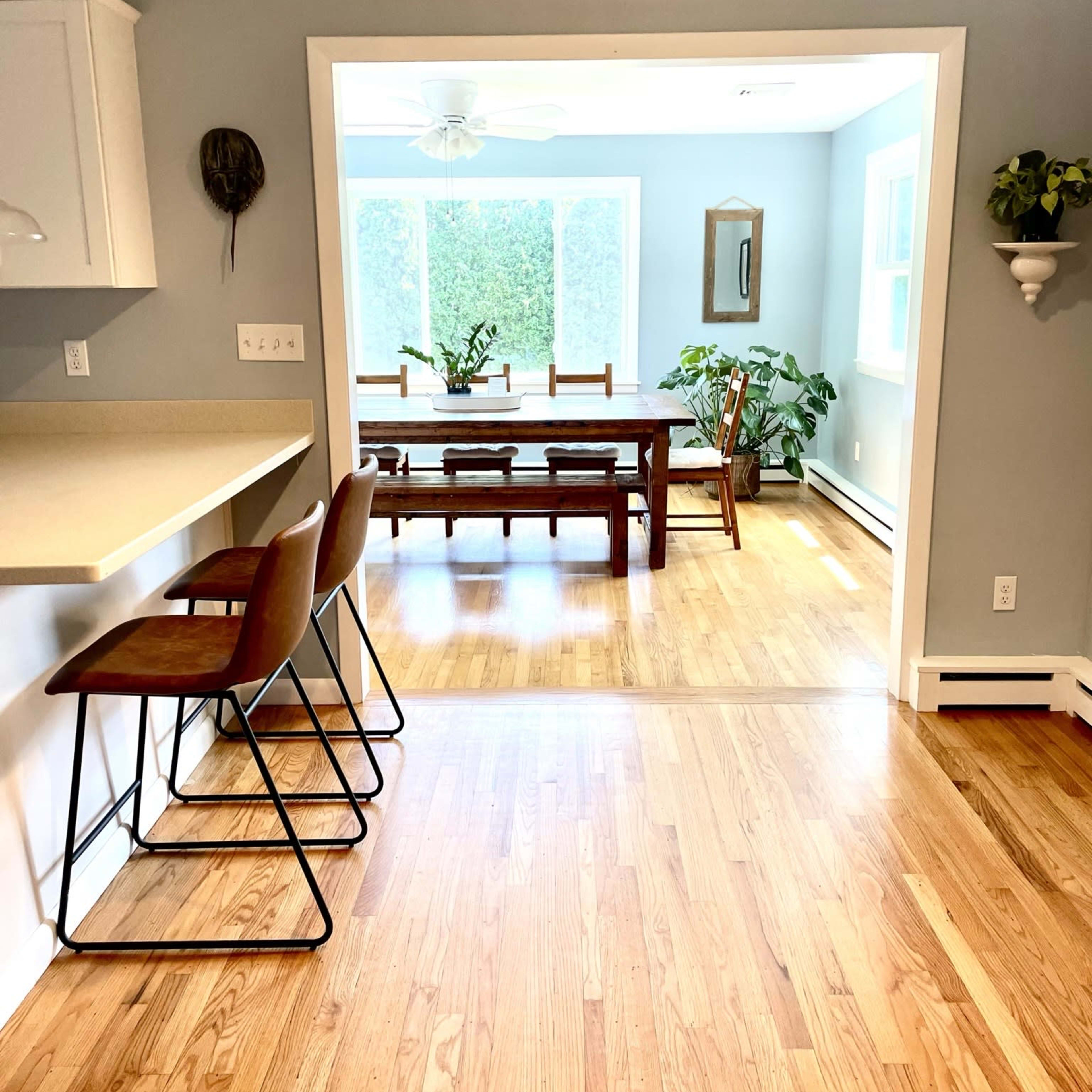 A kitchen with bar stools overlooks a dining area featuring a large wooden table and plants, separated by a doorway.