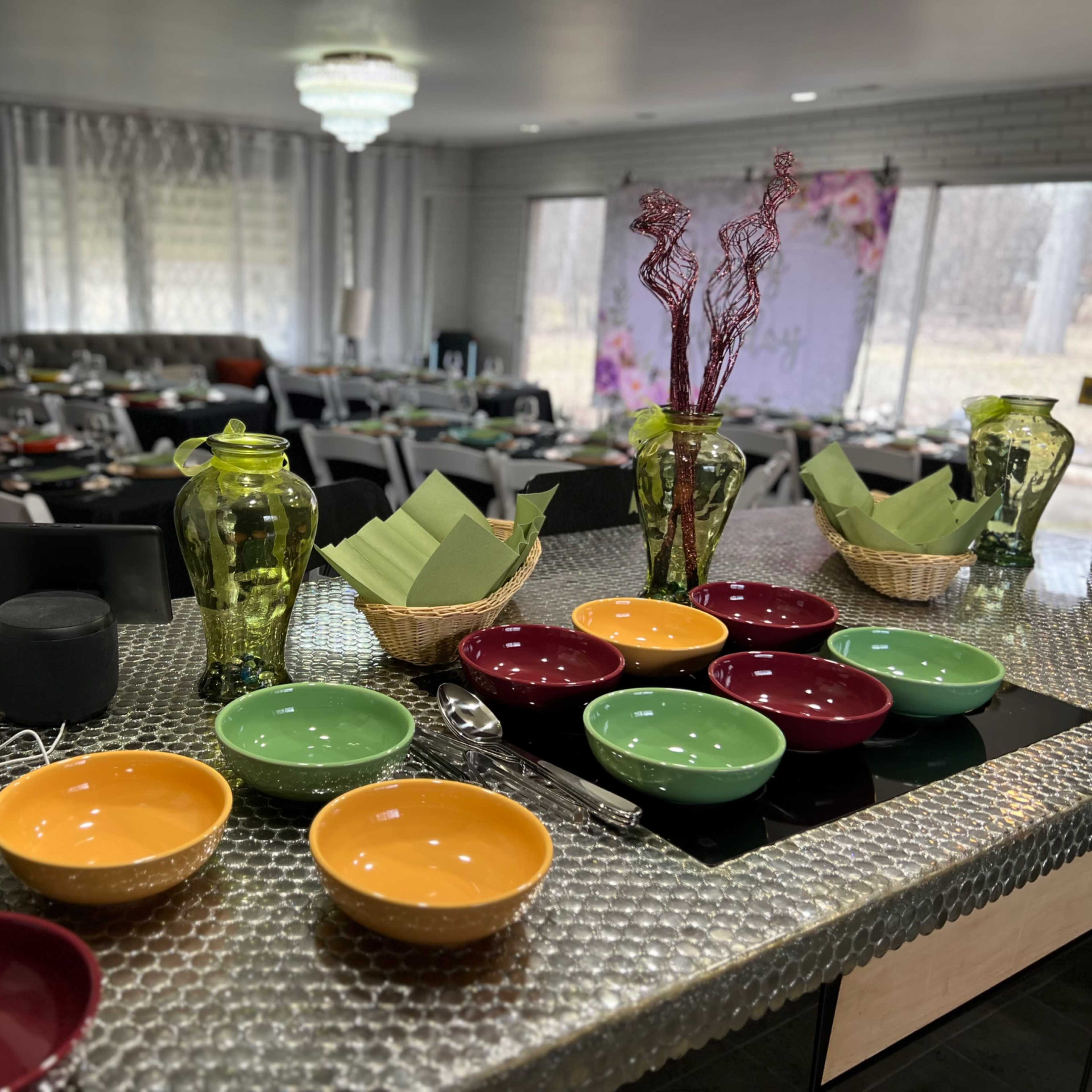 A dining area is set with colorful bowls and decorative items on a countertop, while chairs and tables are arranged for a gathering in the background.