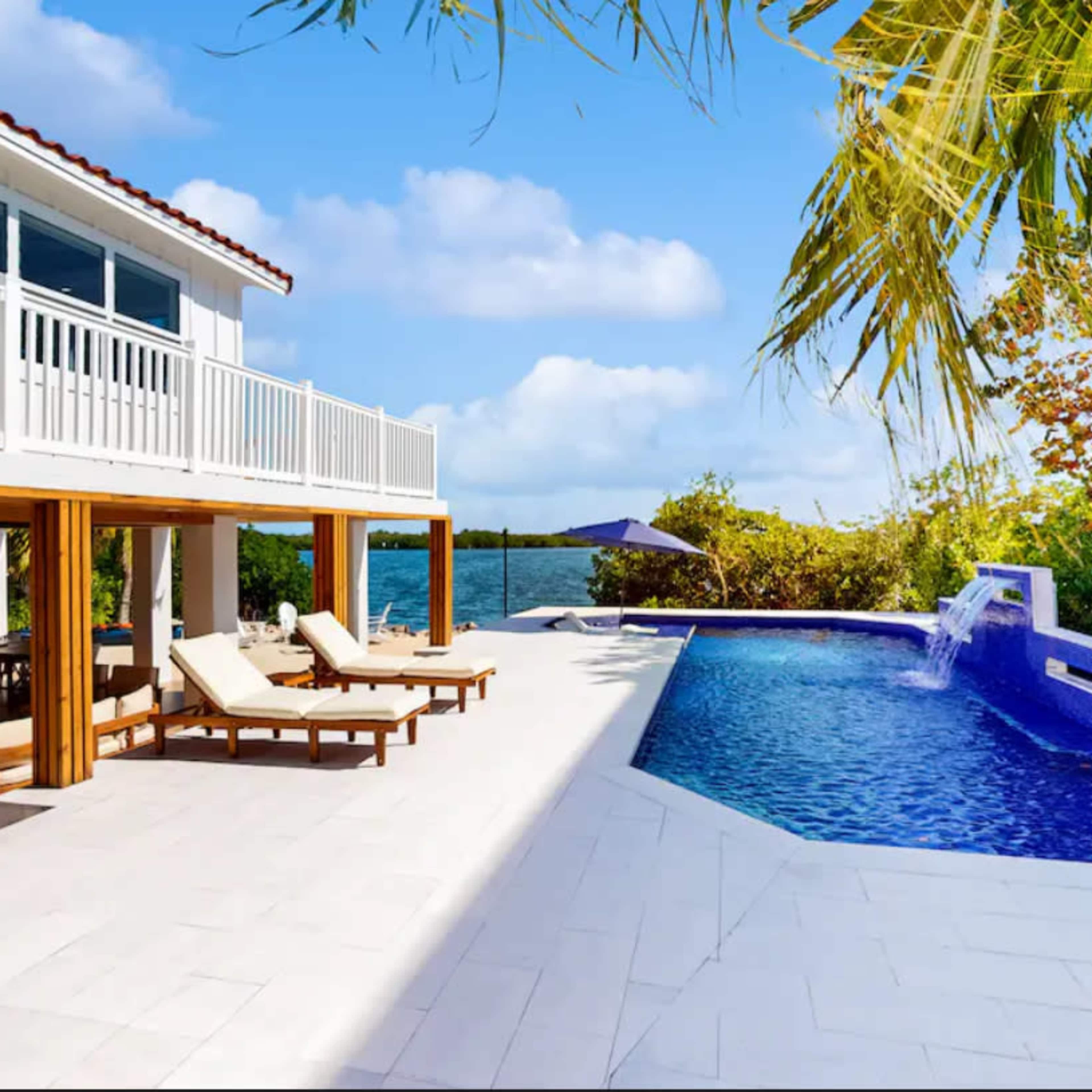 A modern waterfront house features an outdoor patio with lounge chairs and a swimming pool surrounded by tropical vegetation.