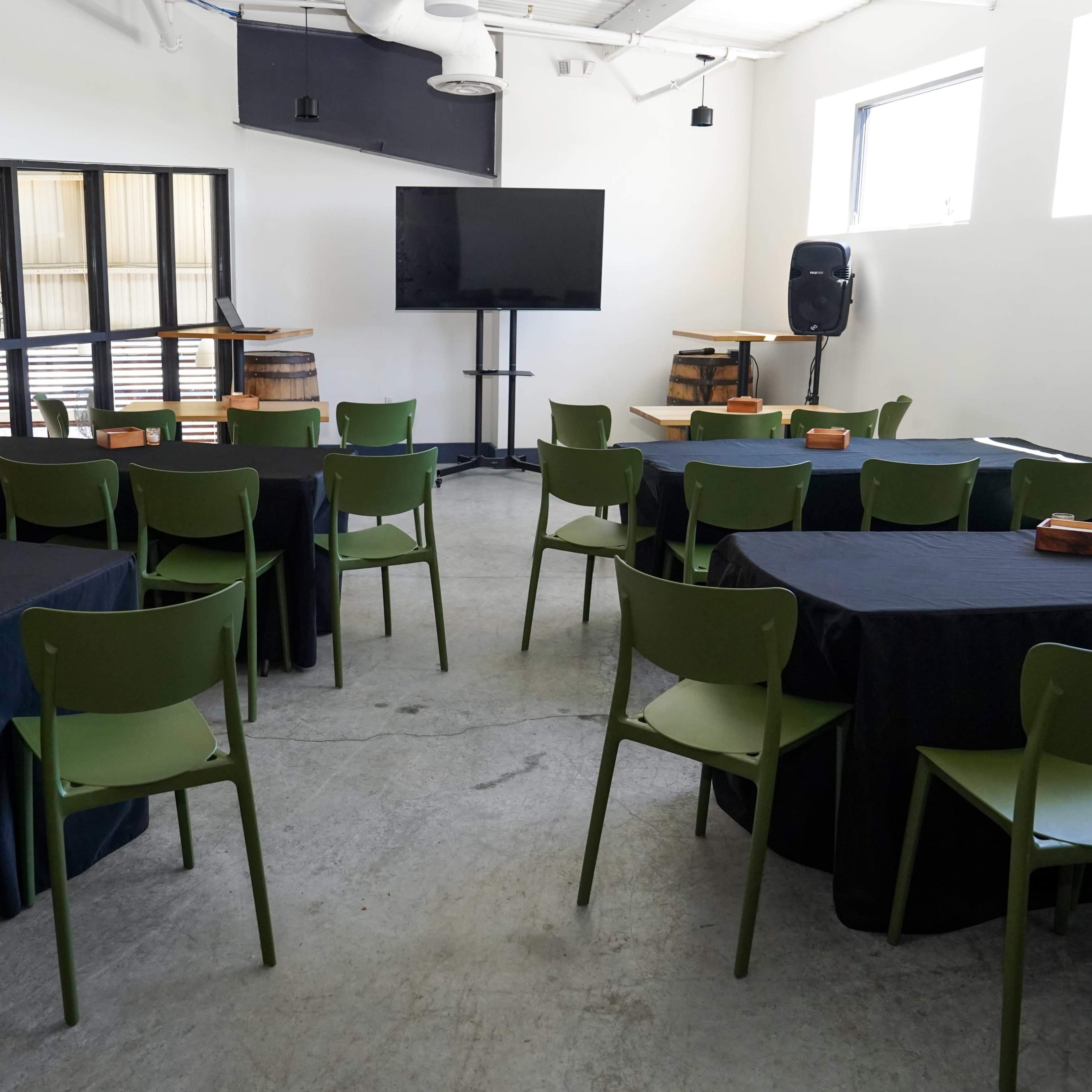 A meeting room with several green chairs around black tables, a large screen on a stand, and large windows providing natural light.