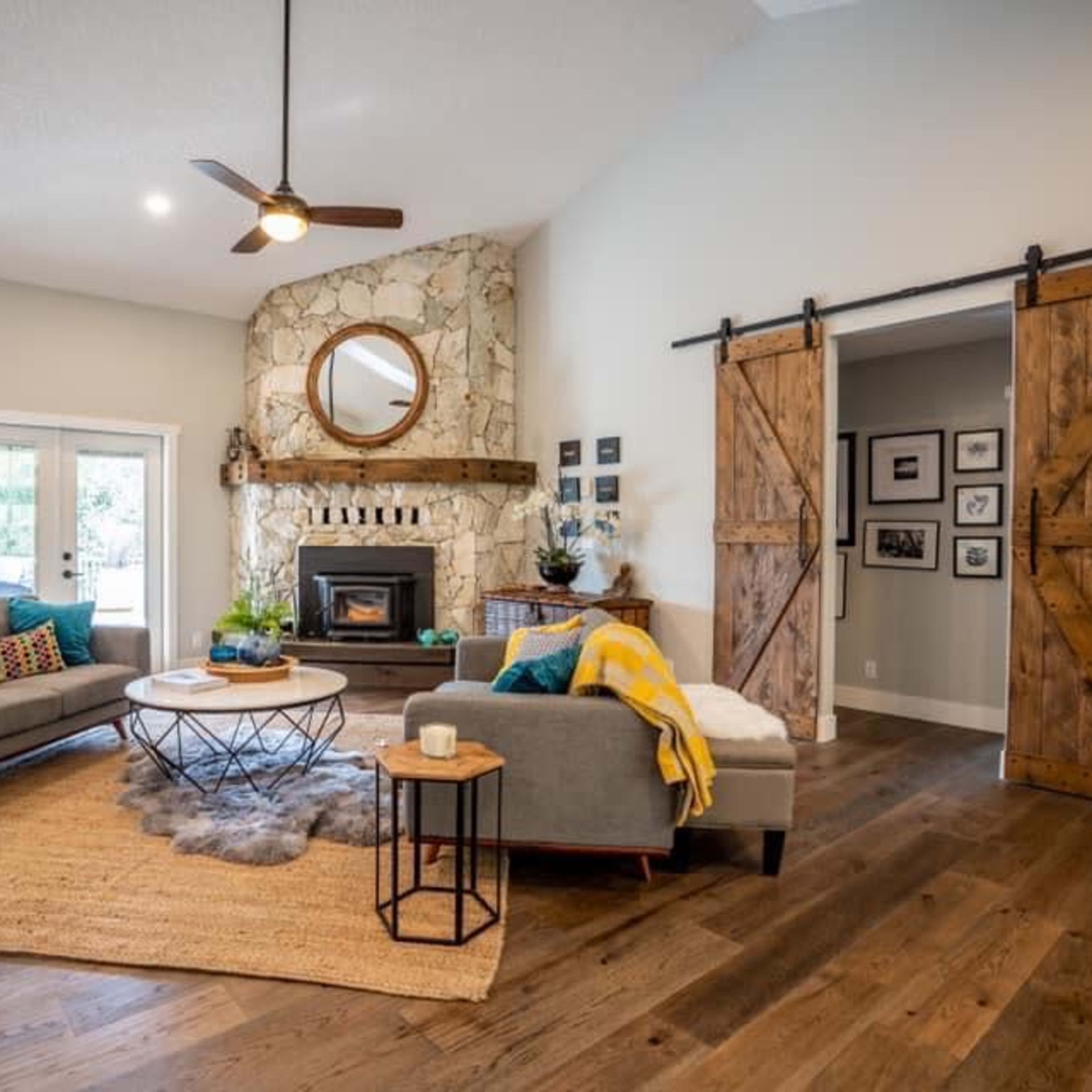A modern living room with two gray sofas, a circular coffee table, a stone fireplace with a round mirror above it, and sliding barn-style doors, all set on a wooden floor.