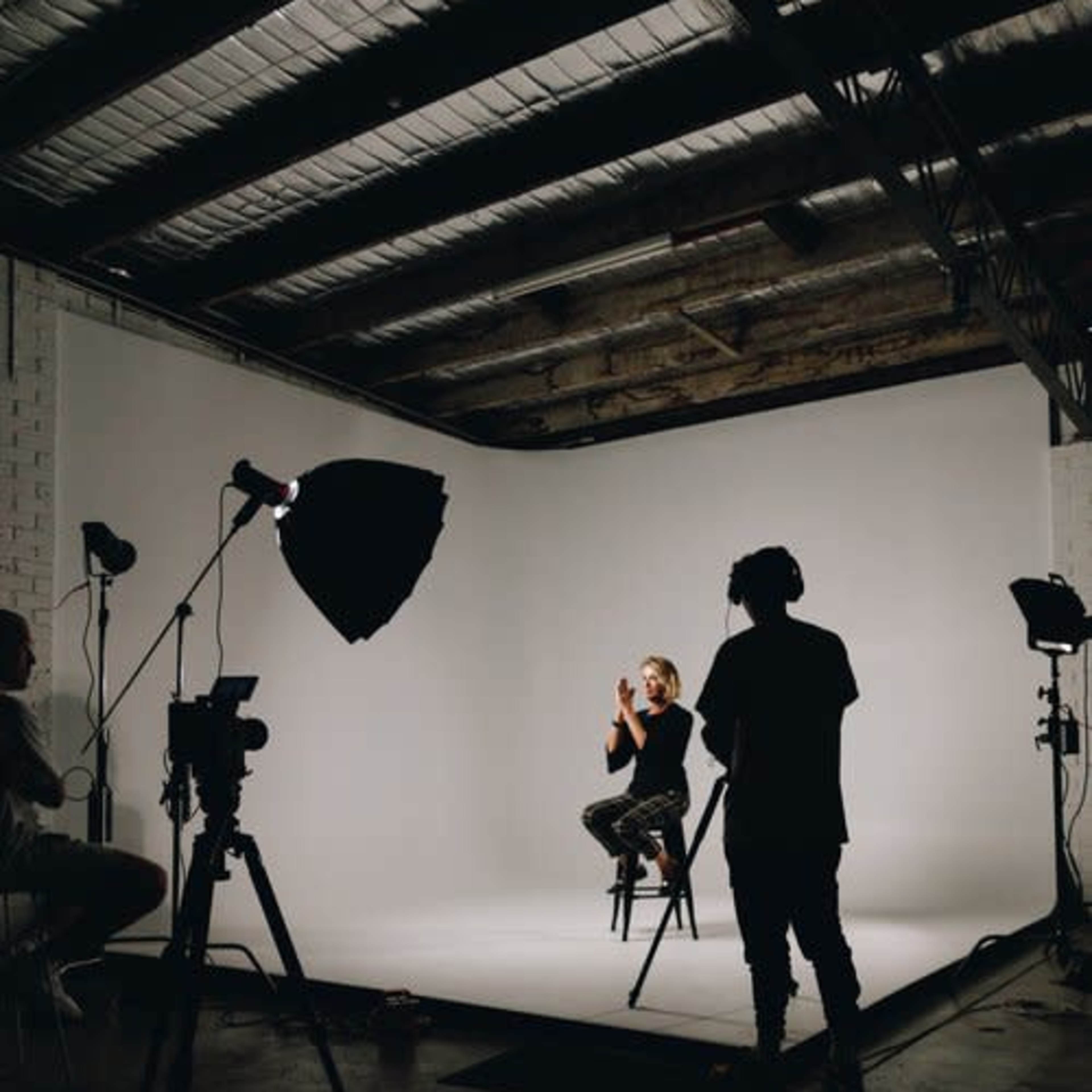 A person sits on a stool in a studio setting, illuminated by multiple lights and surrounded by cameras on tripods.