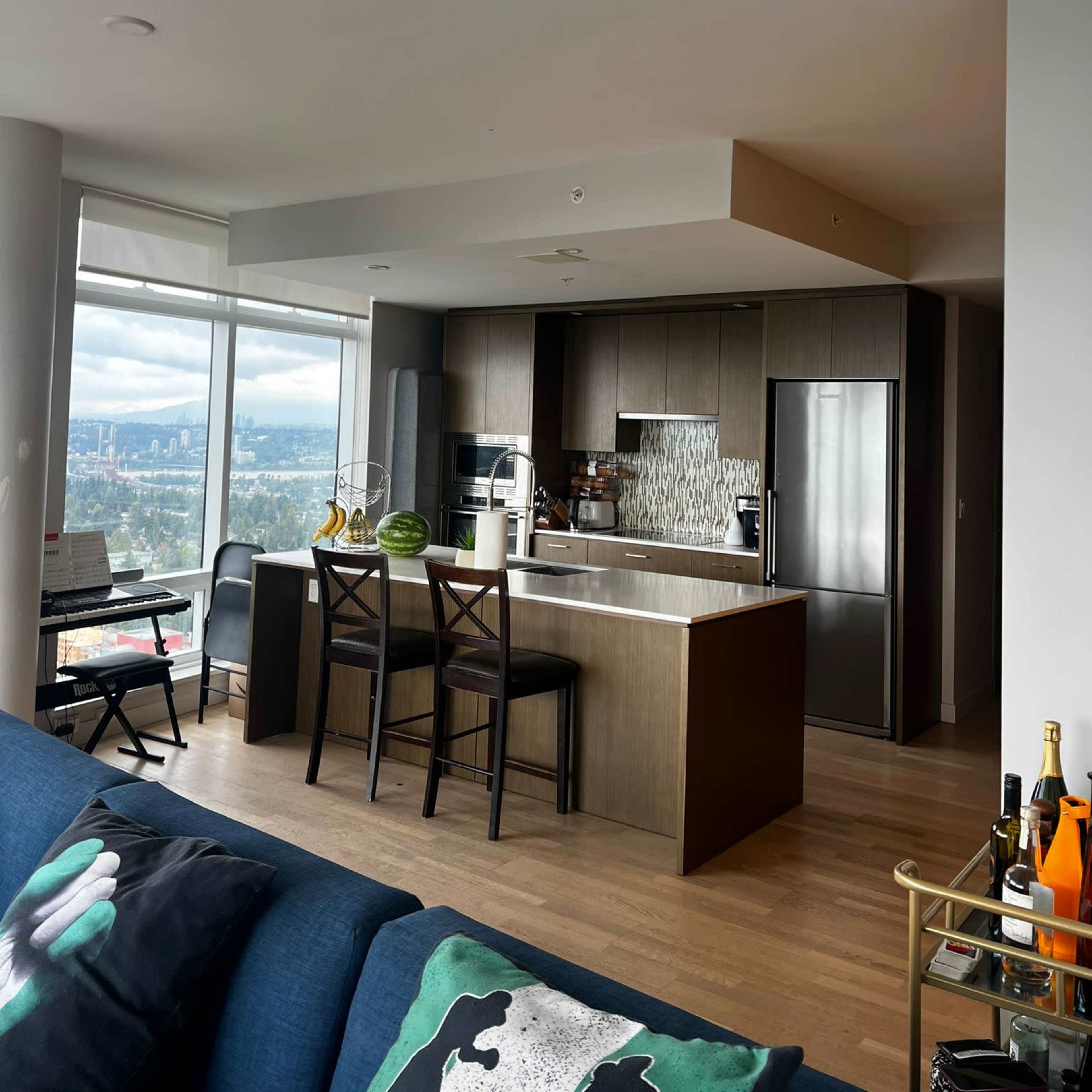 A modern kitchen features dark cabinetry, a large island, and a view of the city through a floor-to-ceiling window.