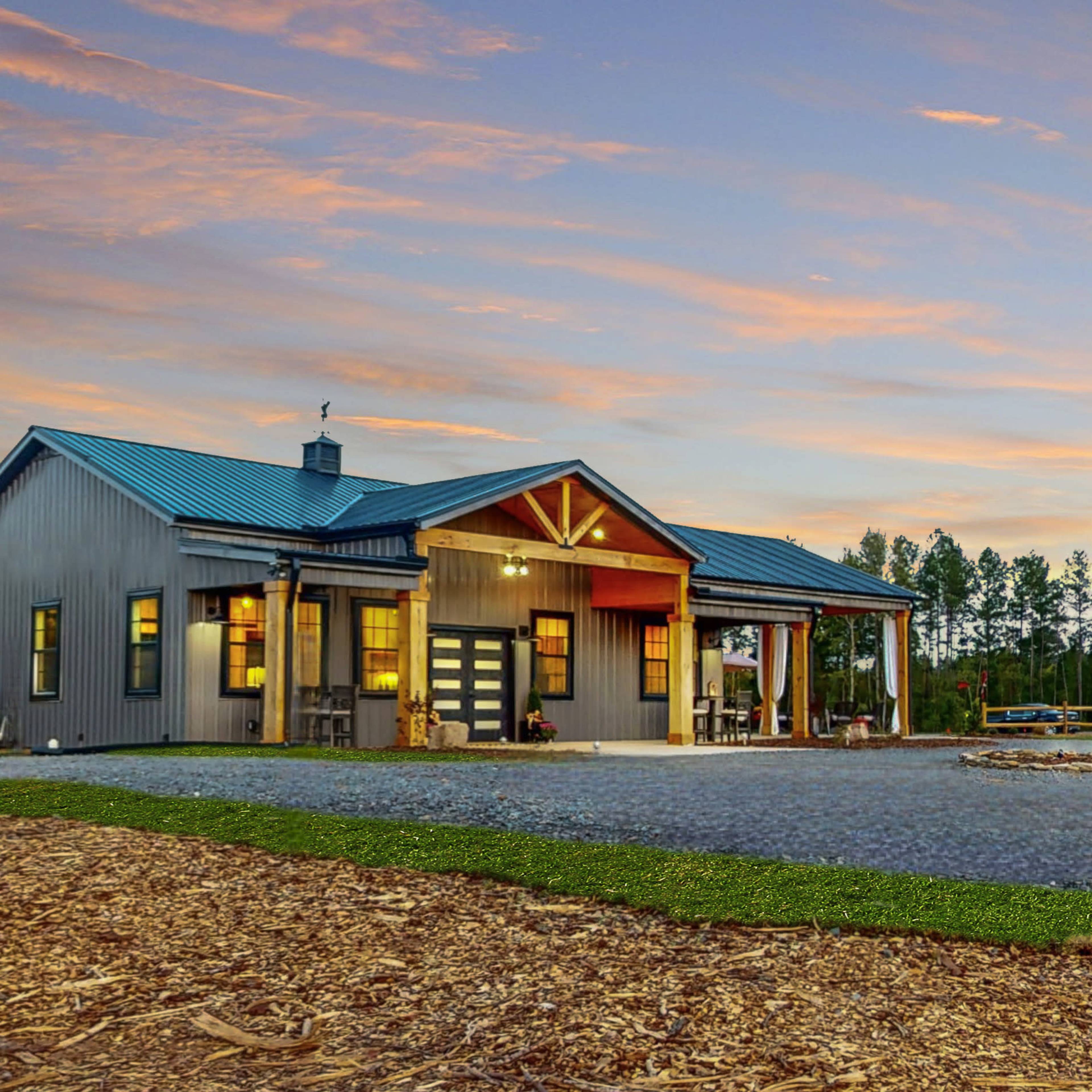 A modern house with a metal roof and wooden accents is pictured beside a circular fire pit area surrounded by trees at sunset.