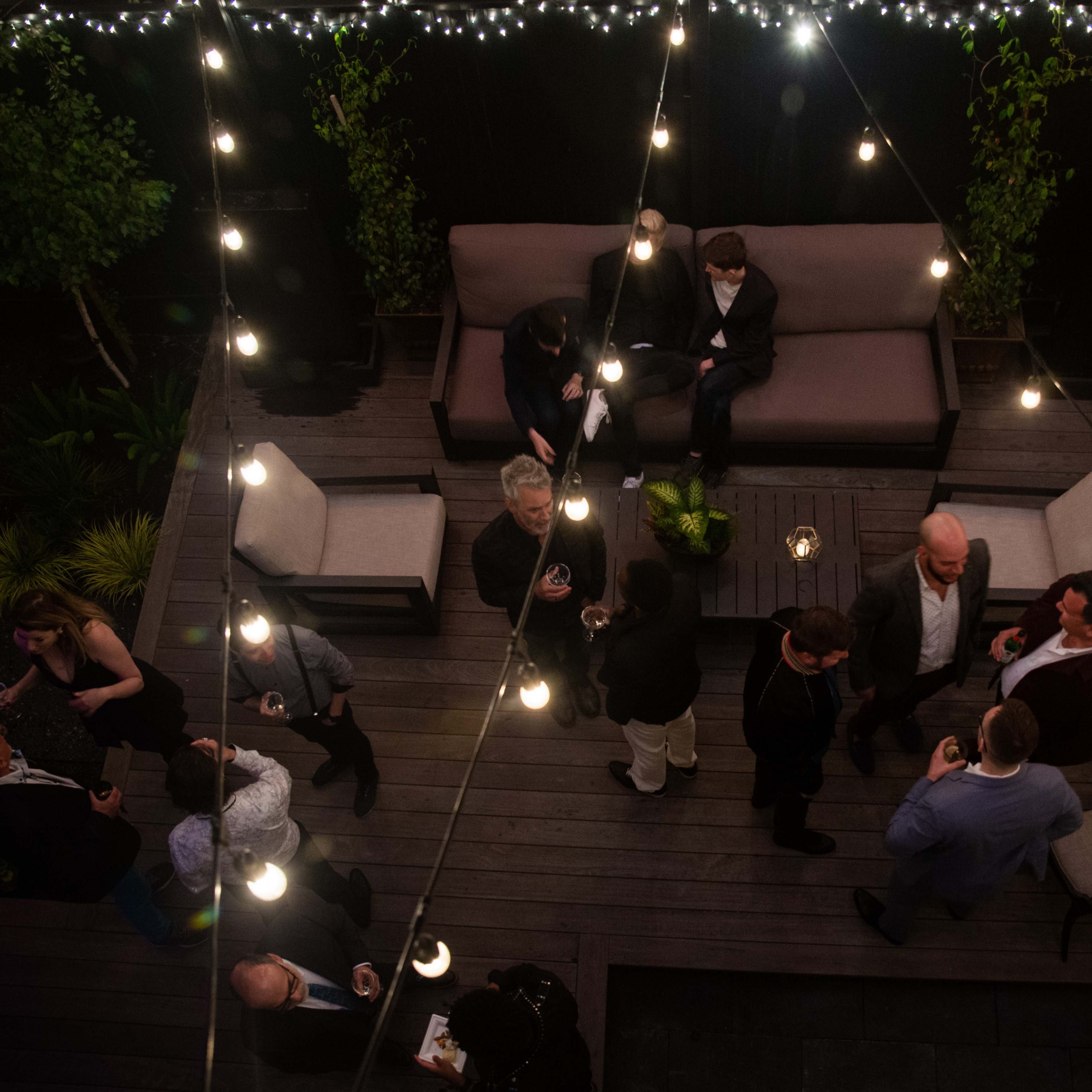 A rooftop gathering features people in formal attire conversing and socializing under string lights.