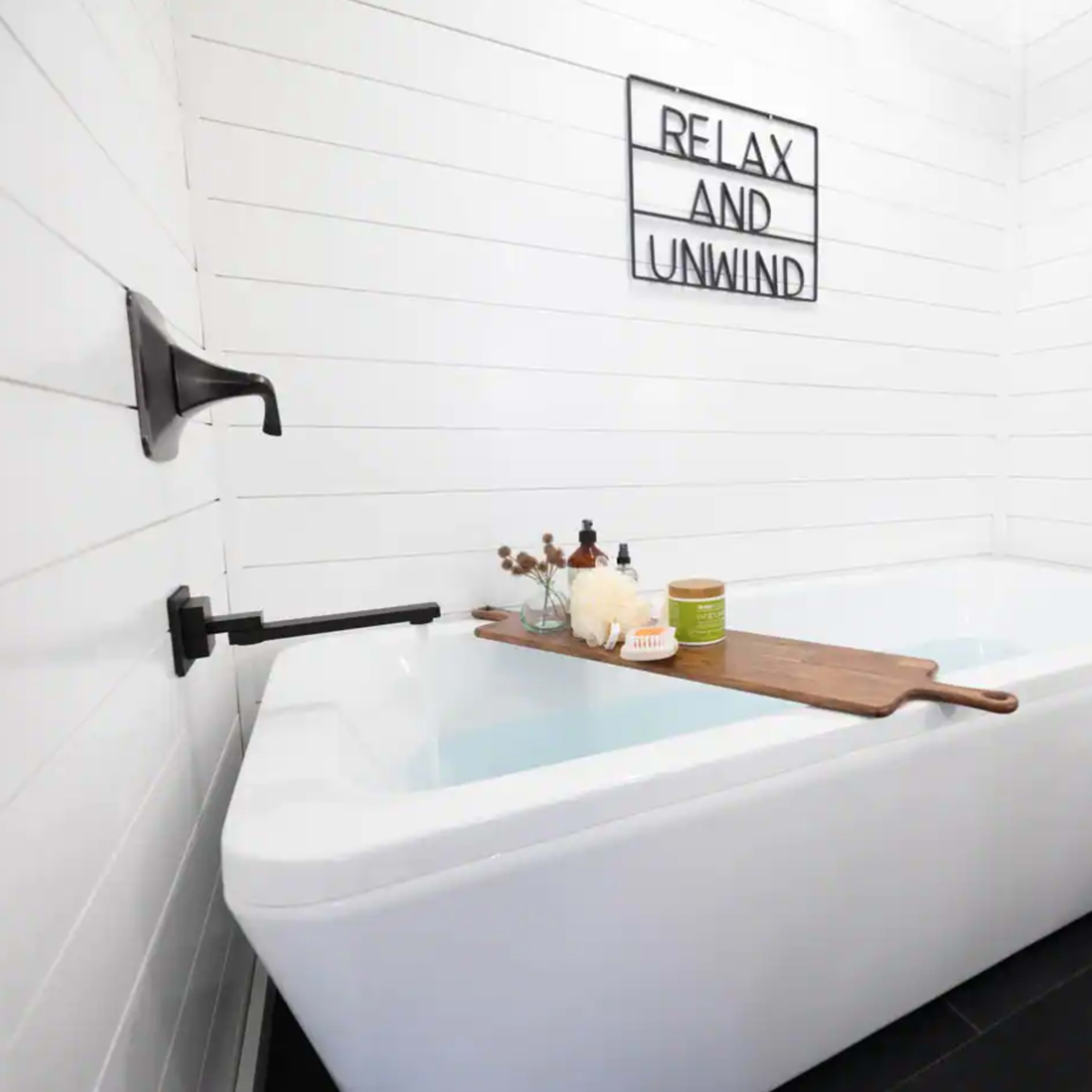 A modern bathtub with a wooden bath tray rests against white shiplap walls, featuring a sign that reads "RELAX AND UNWIND."