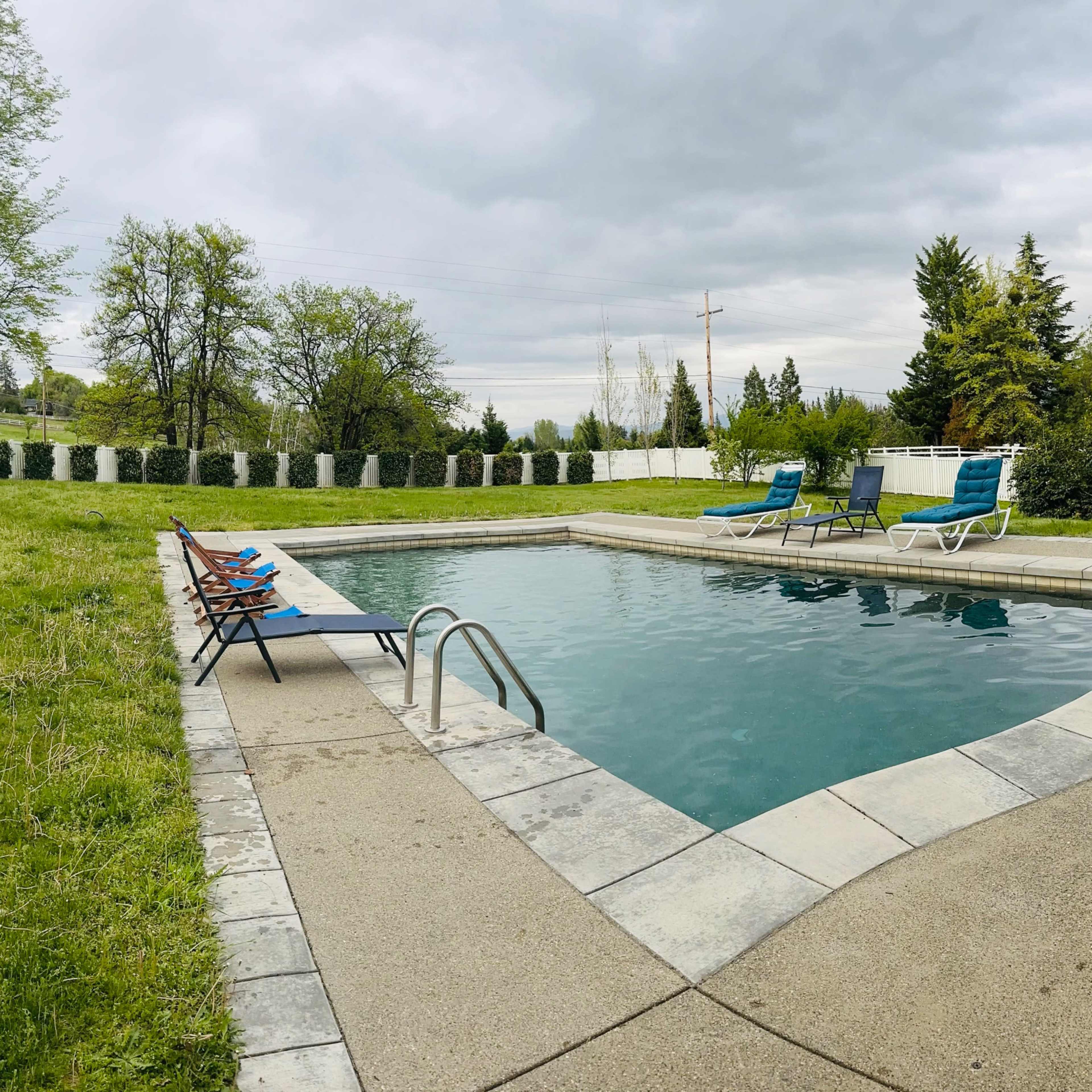 A rectangular swimming pool surrounded by a grassy area and several lounge chairs under a cloudy sky.