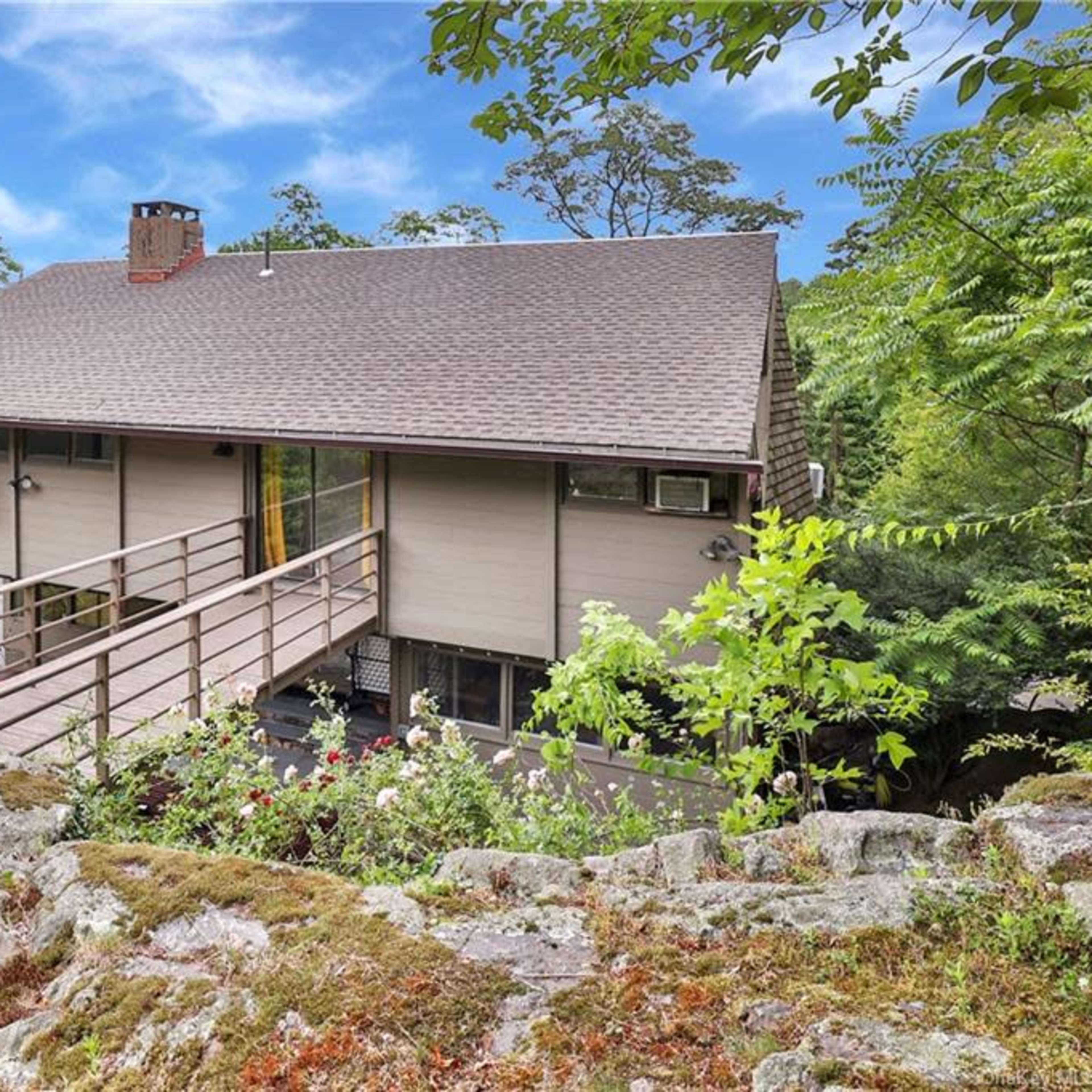 A modern two-story house sits on a rocky hillside, surrounded by trees and greenery.