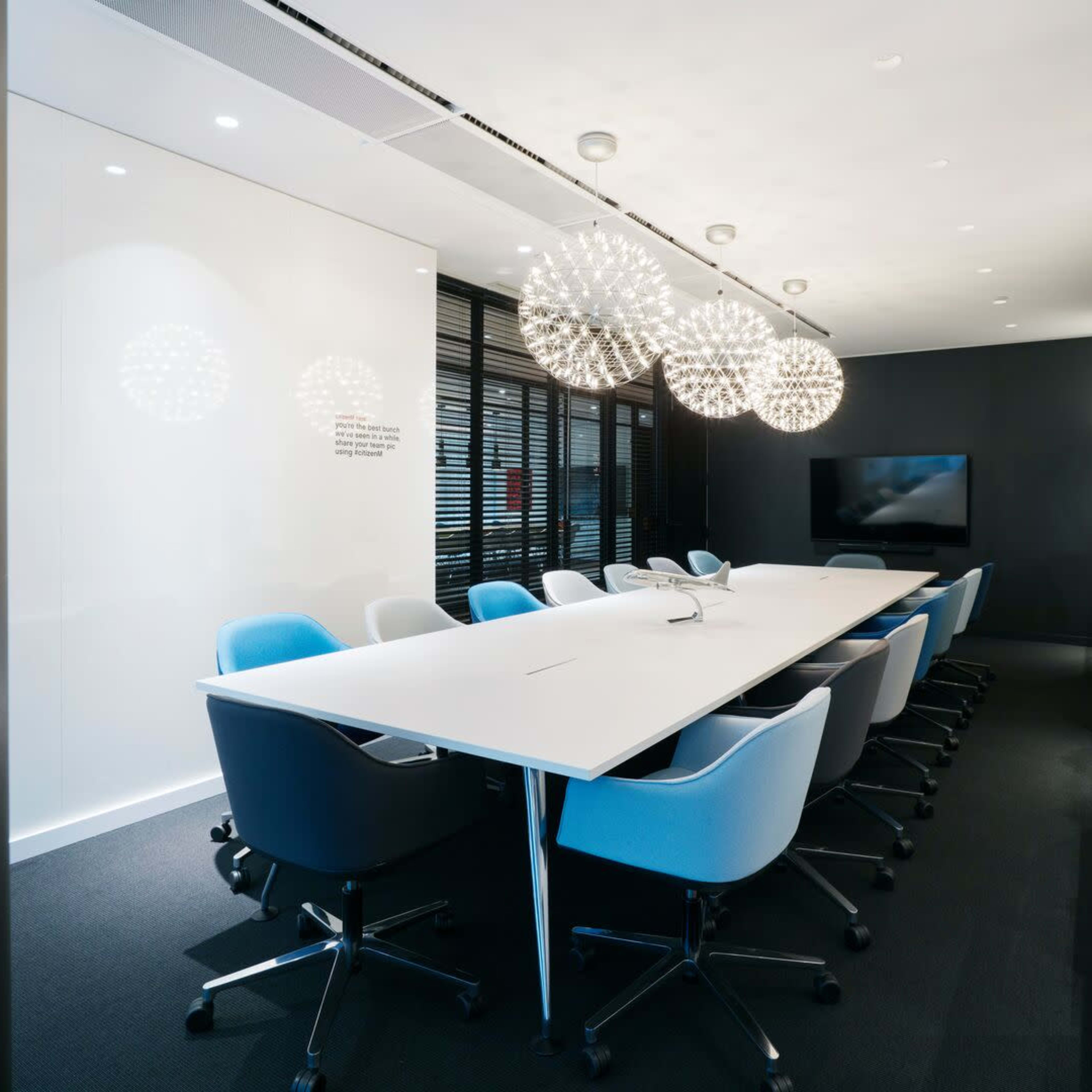 A modern conference room features a long white table surrounded by blue and gray office chairs, with large spherical light fixtures overhead and a wall-mounted screen at one end.