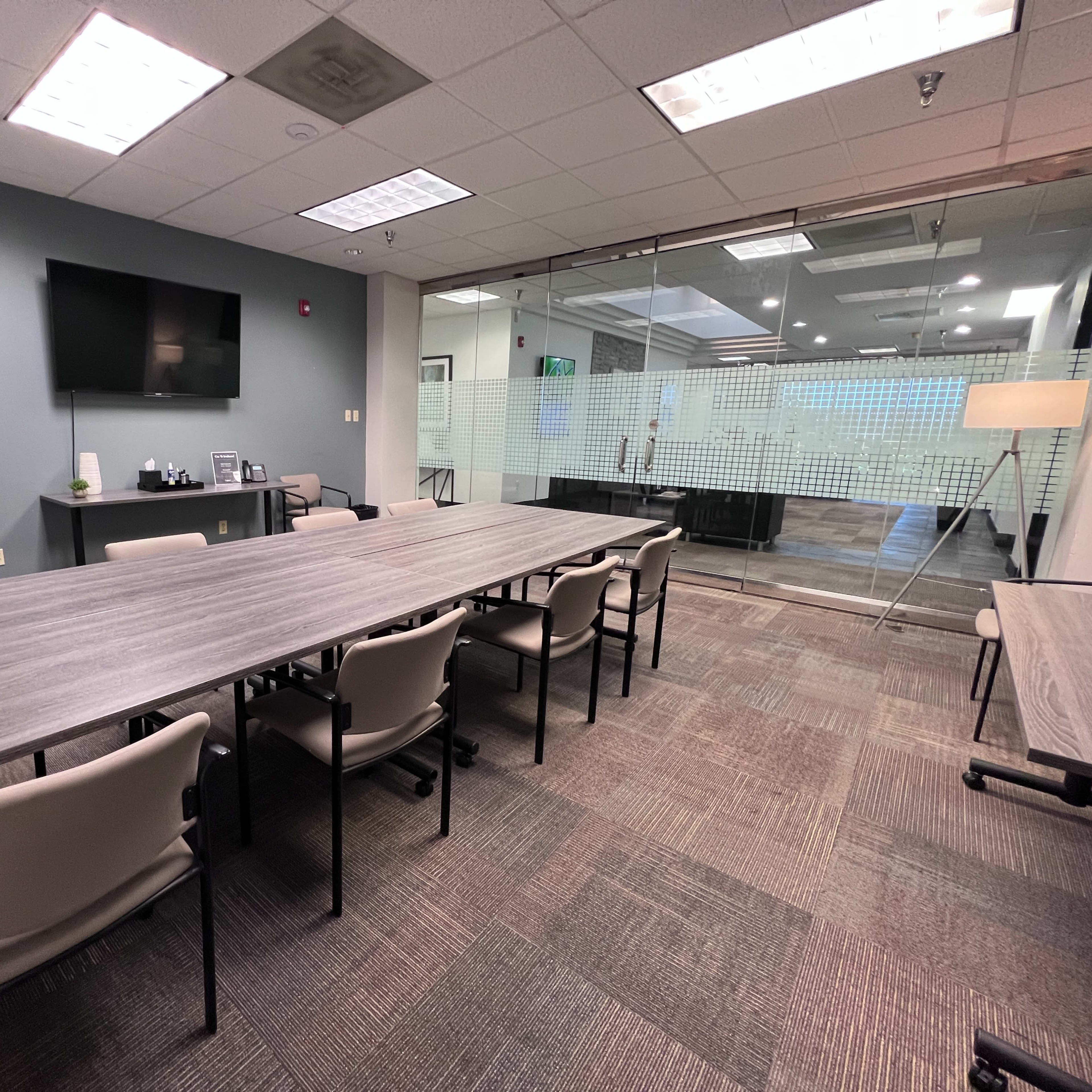 A meeting room with a long table, several chairs, a television, and glass walls leading to another area.