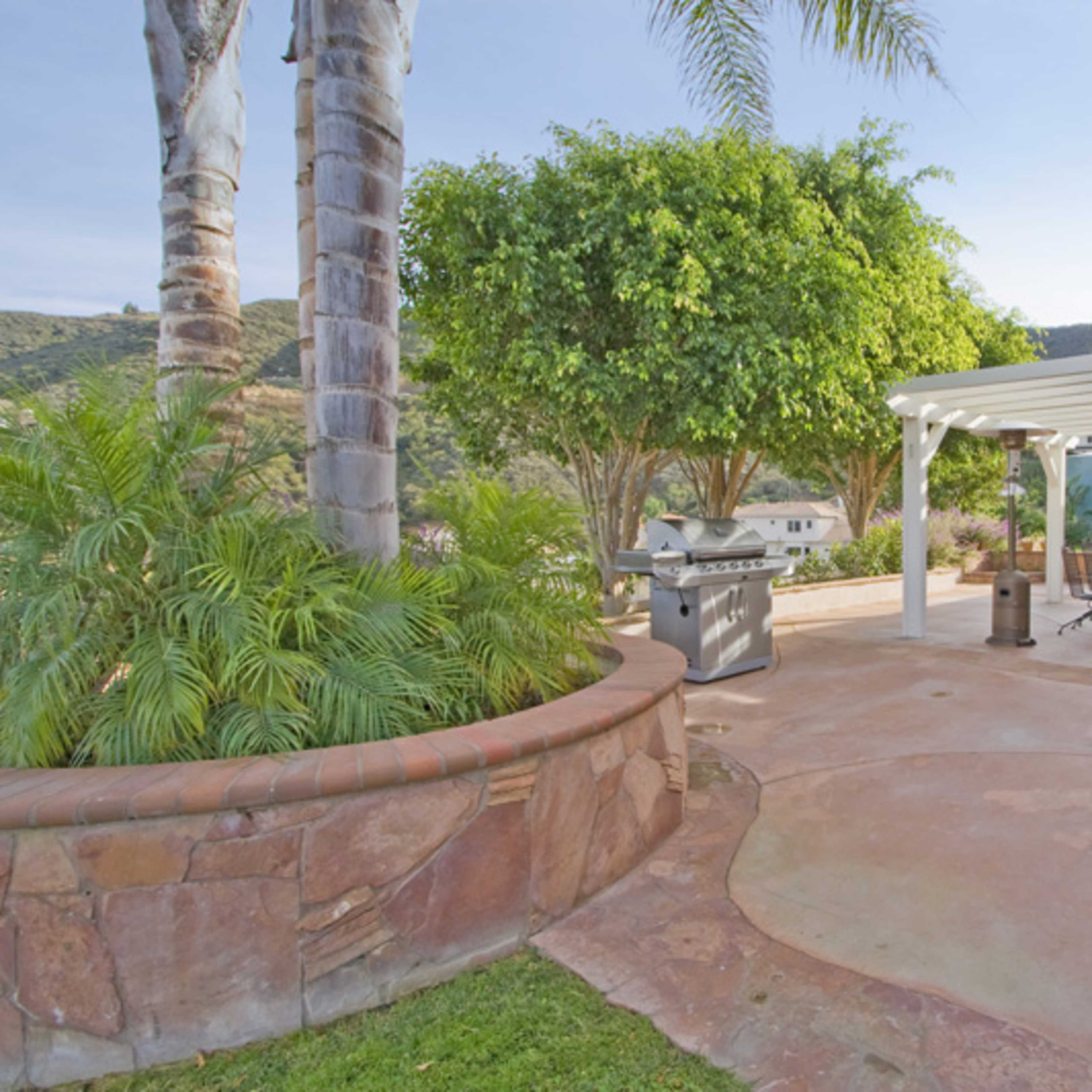 A patio area with a grill, table, and chairs surrounded by greenery and hills in the background.