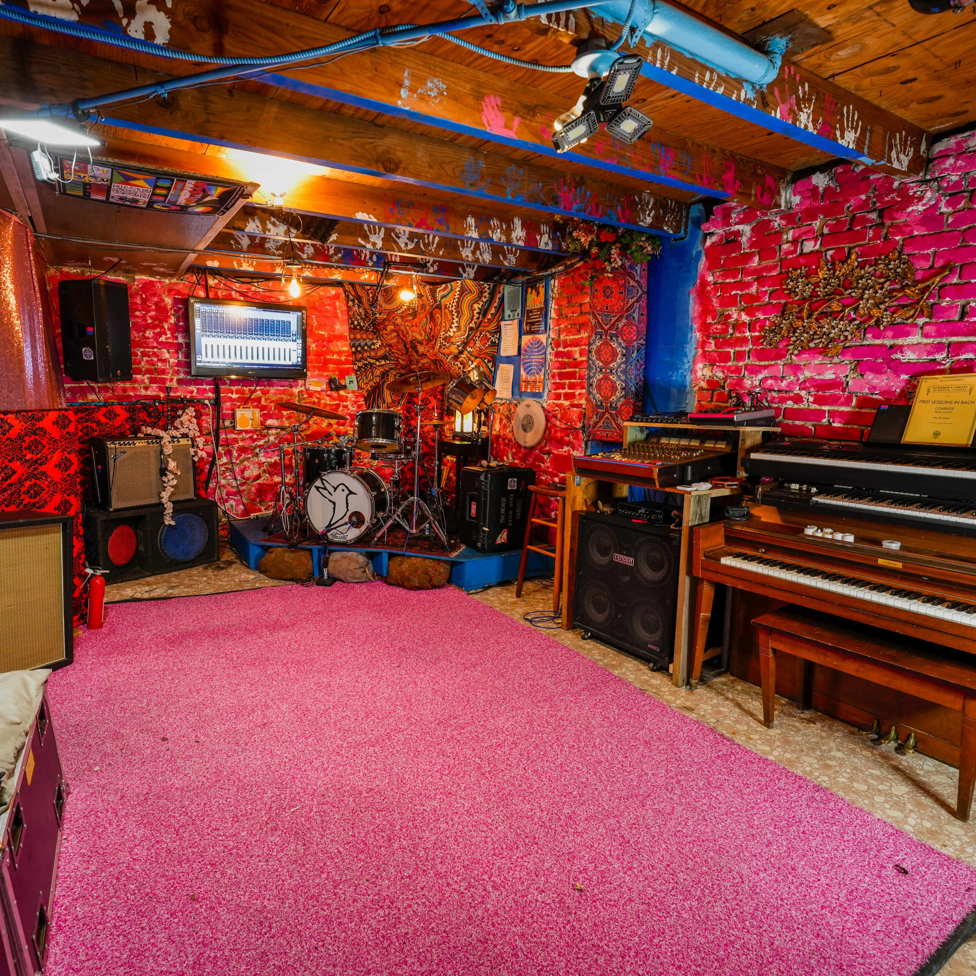 A colorful basement room featuring a drum set, a piano, and various amplifiers, all arranged around a pink carpet.