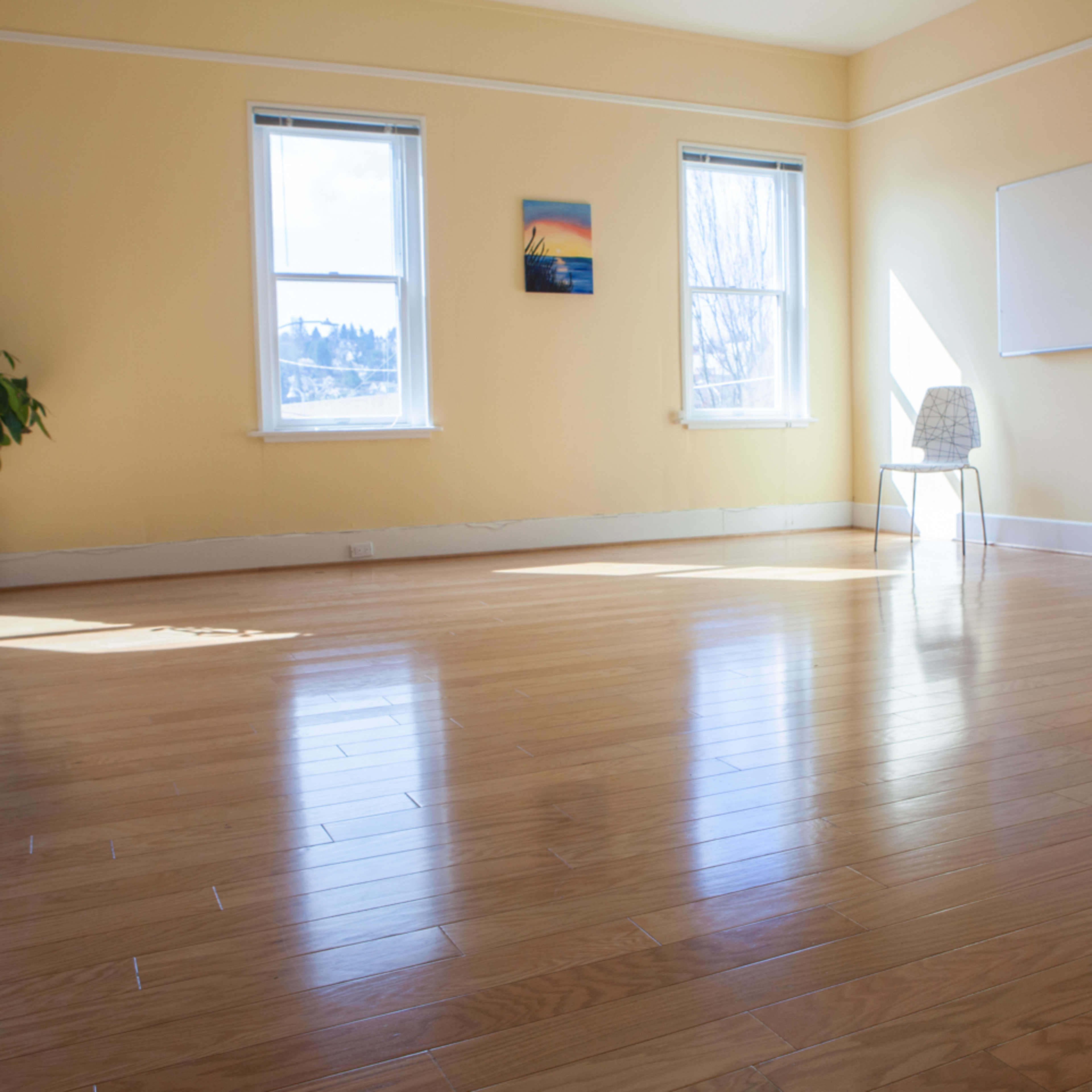 A sunlit room with yellow walls, hardwood floors, two windows, a small table, a chair, and plants in pots.