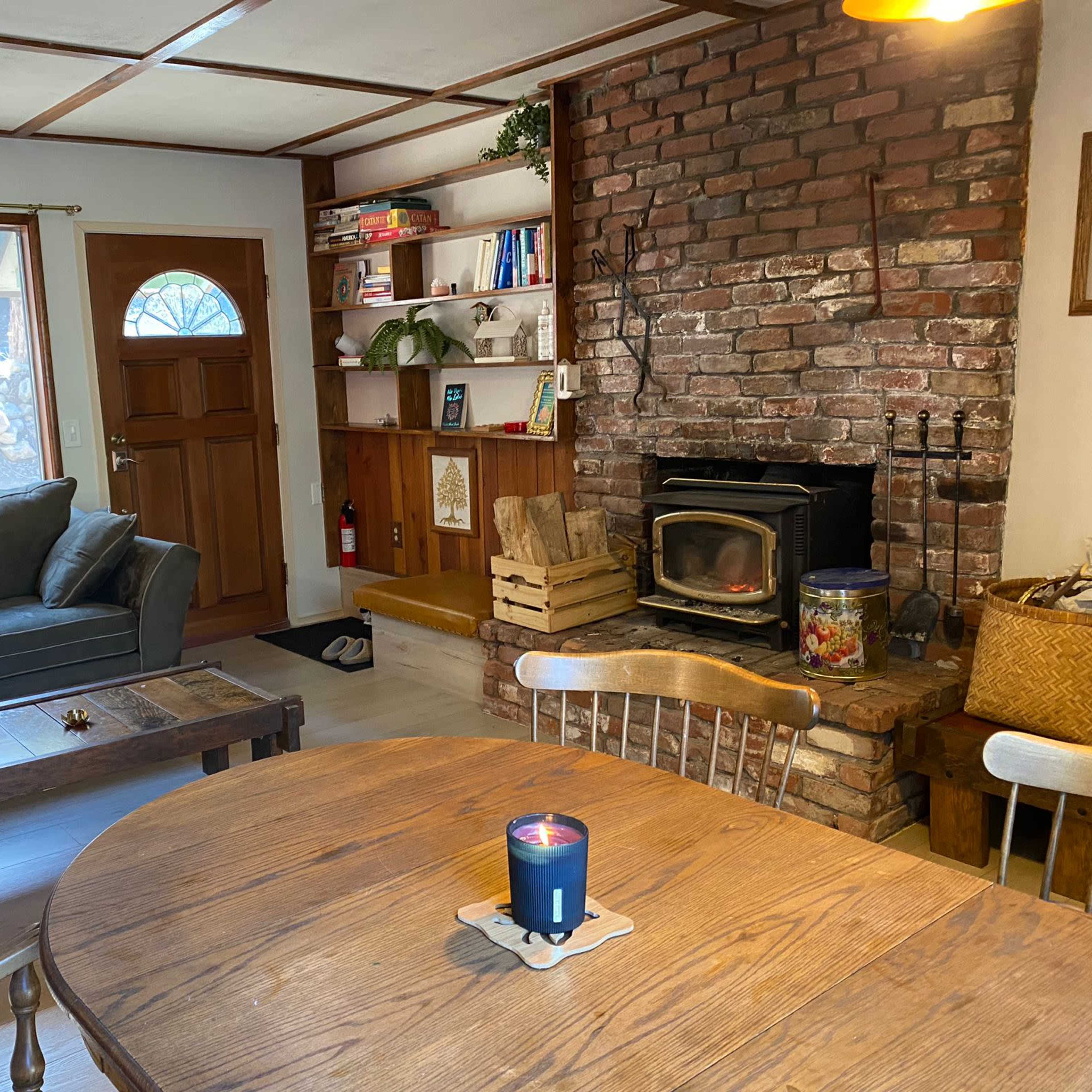 A cozy living room with a brick fireplace, a wooden dining table, a green couch, and a front door leading outside.