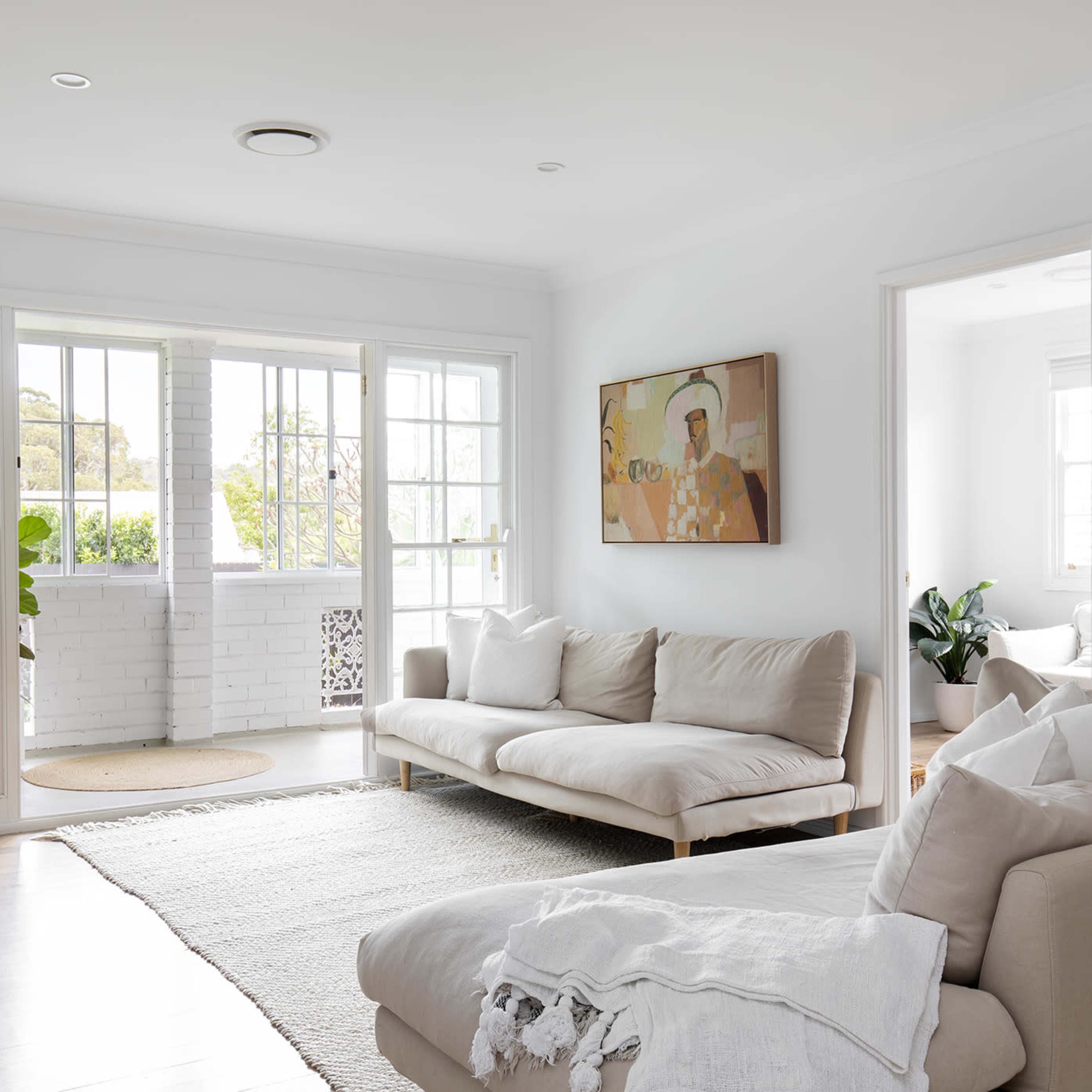 A bright living room with a sofa, a large window, and an open door leading to a patio area, decorated with indoor plants and wall art.
