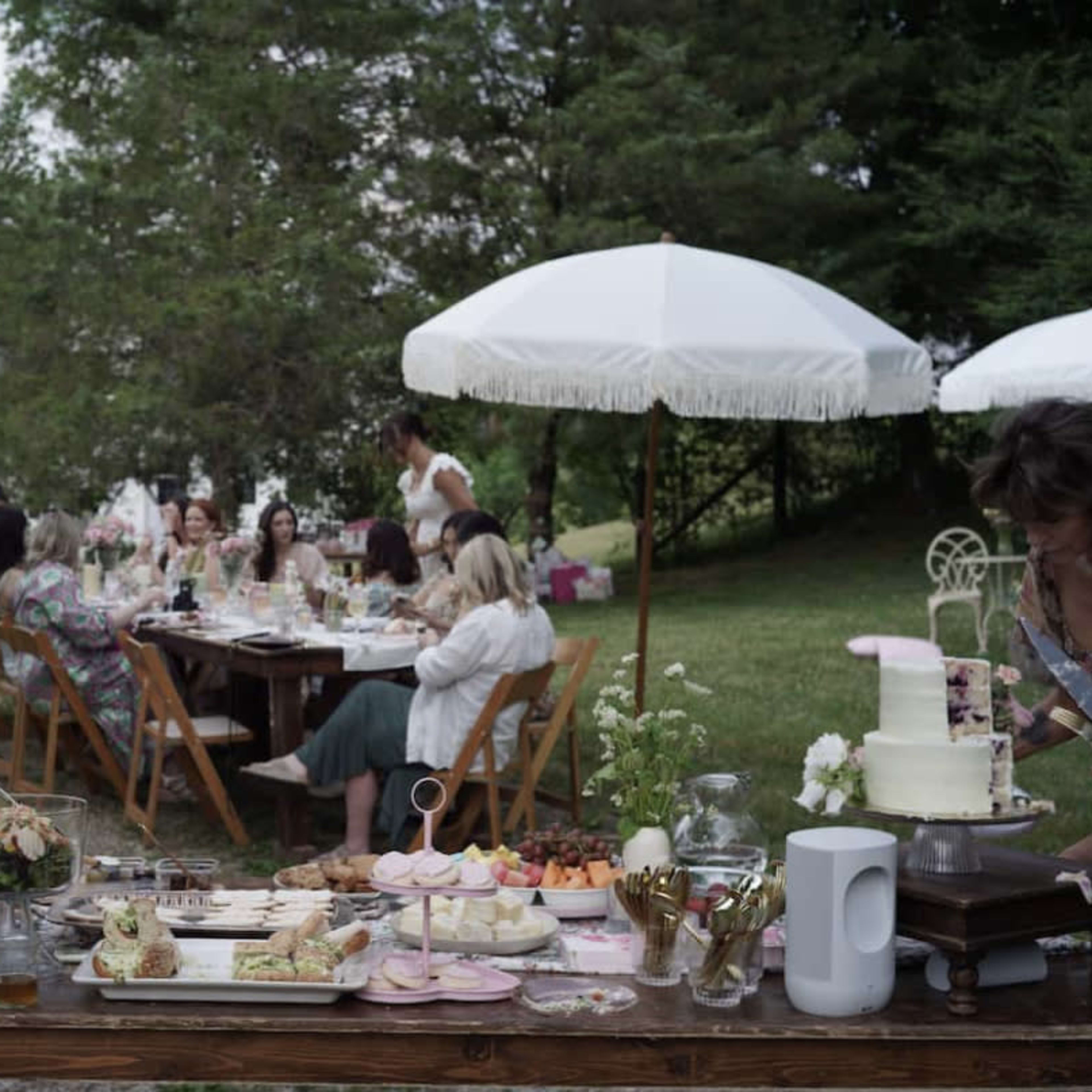 A group of people gathered at a long table outdoors, enjoying a meal under white umbrellas in a garden setting with a house in the background.