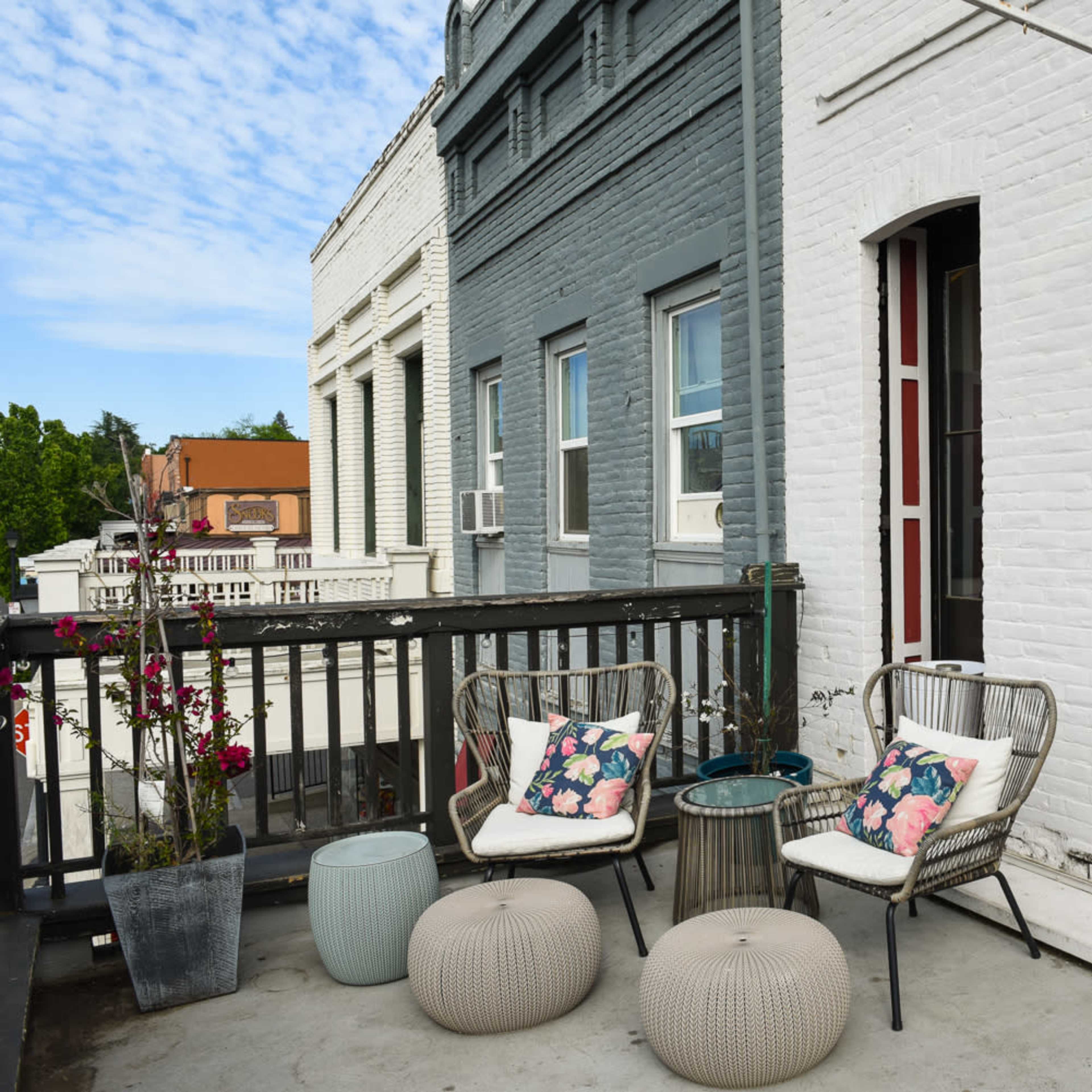 A balcony with two chairs and two poufs, surrounded by potted plants and buildings in the background.