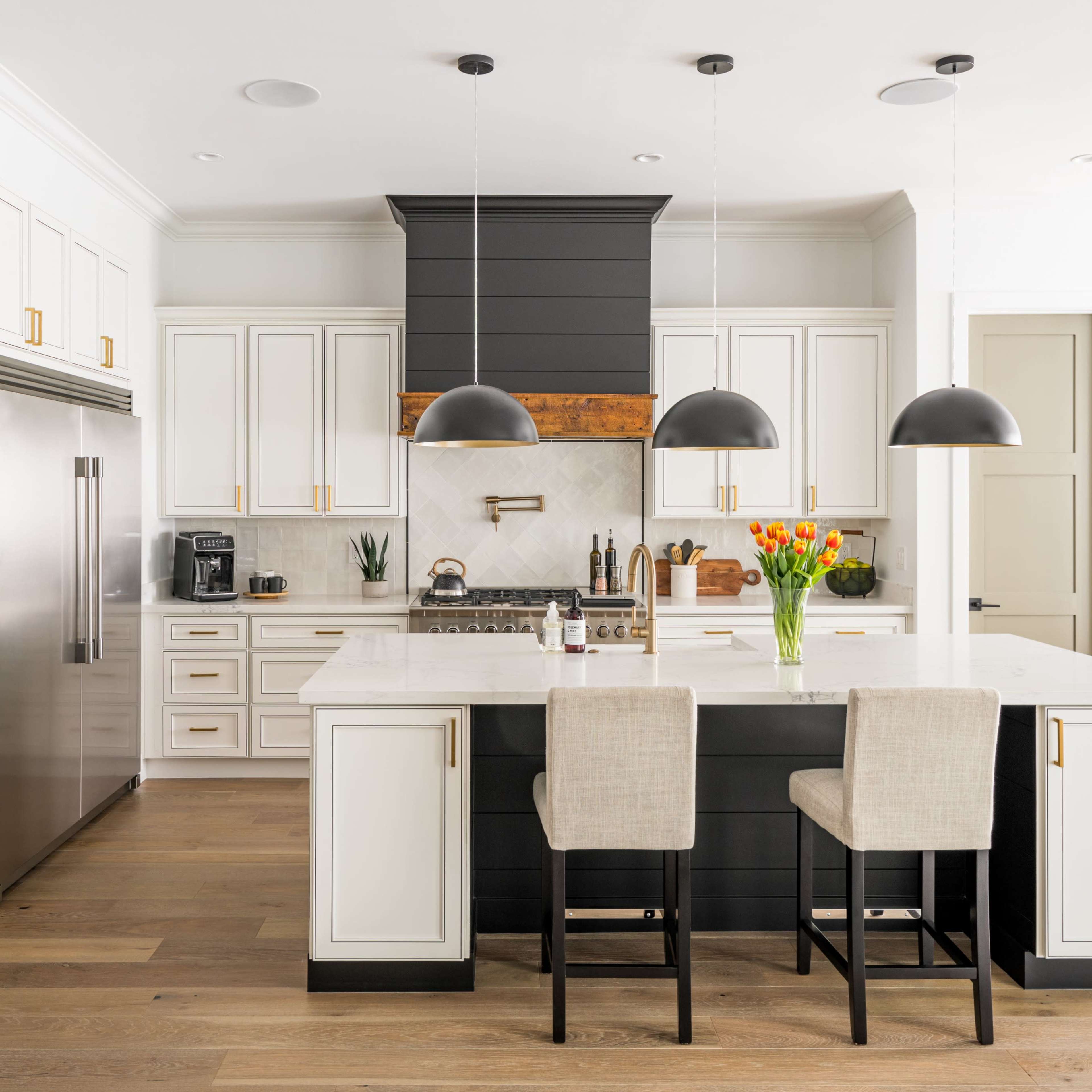 A modern kitchen features white cabinetry, a dark island with two bar stools, granite countertops, and black pendant lights.