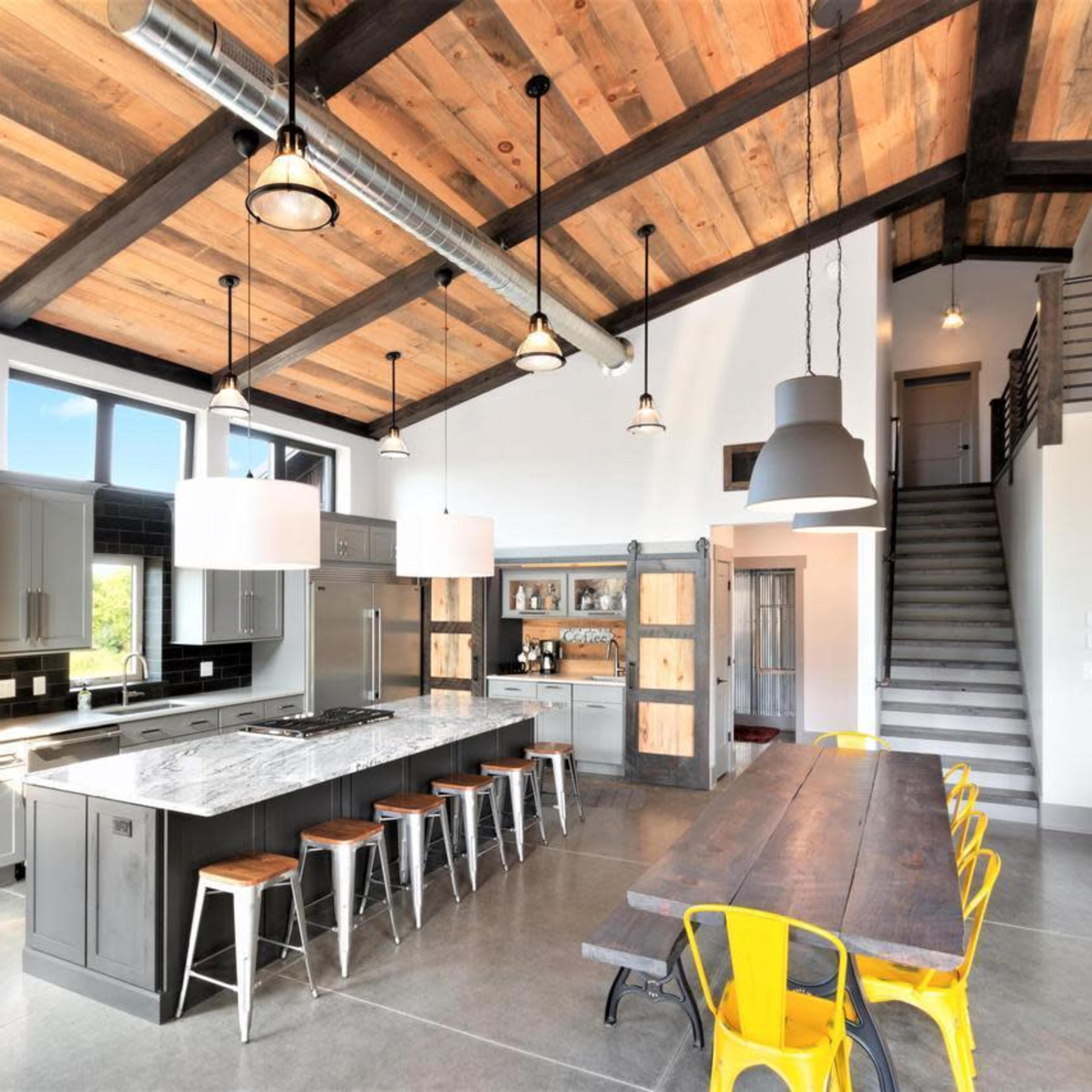 A modern kitchen and dining area with a high wooden ceiling, featuring a large island with a marble countertop, stainless steel appliances, and a long dining table surrounded by yellow chairs.