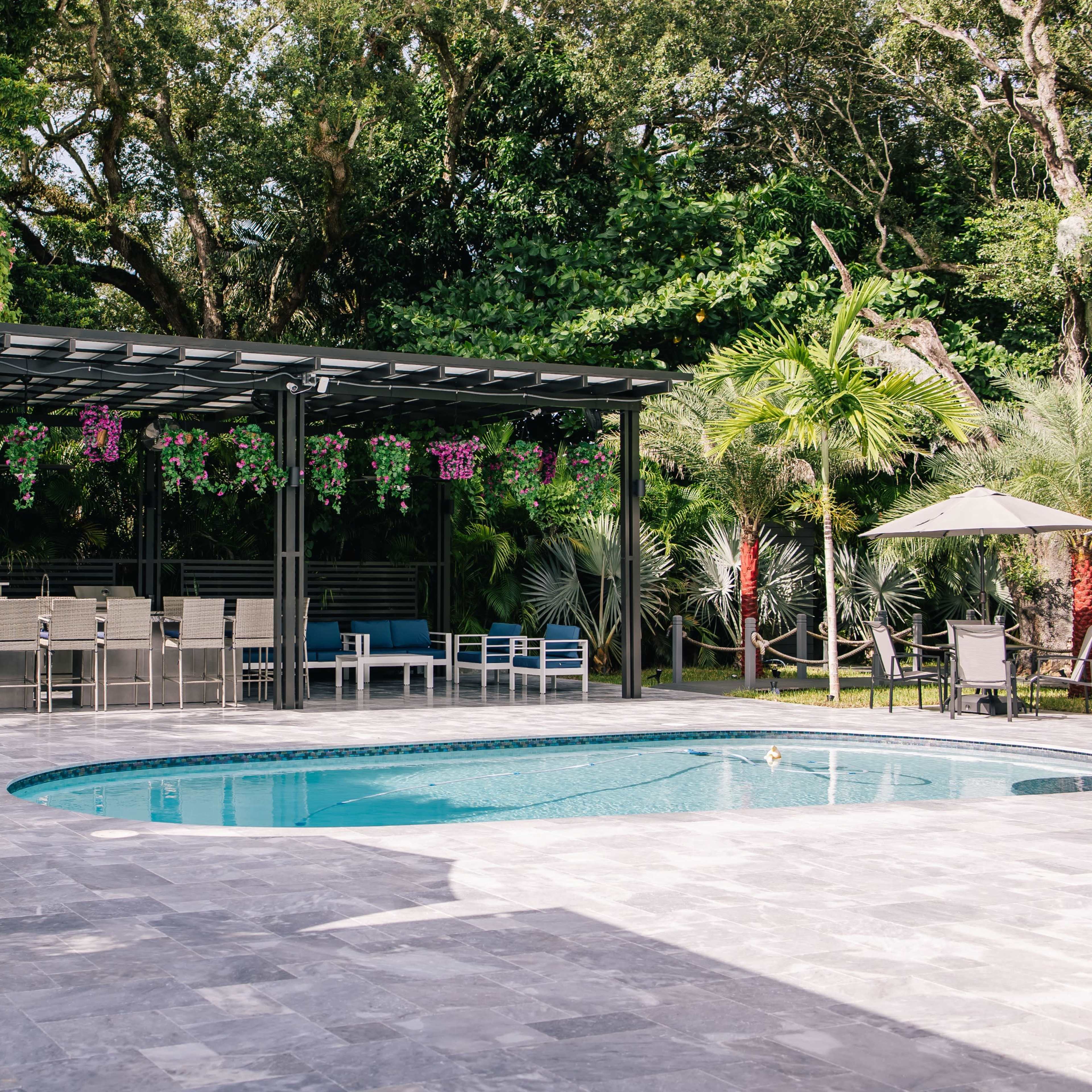 A landscaped outdoor area featuring a swimming pool surrounded by lounge chairs and a shaded seating area supported by a pergola adorned with hanging plants.