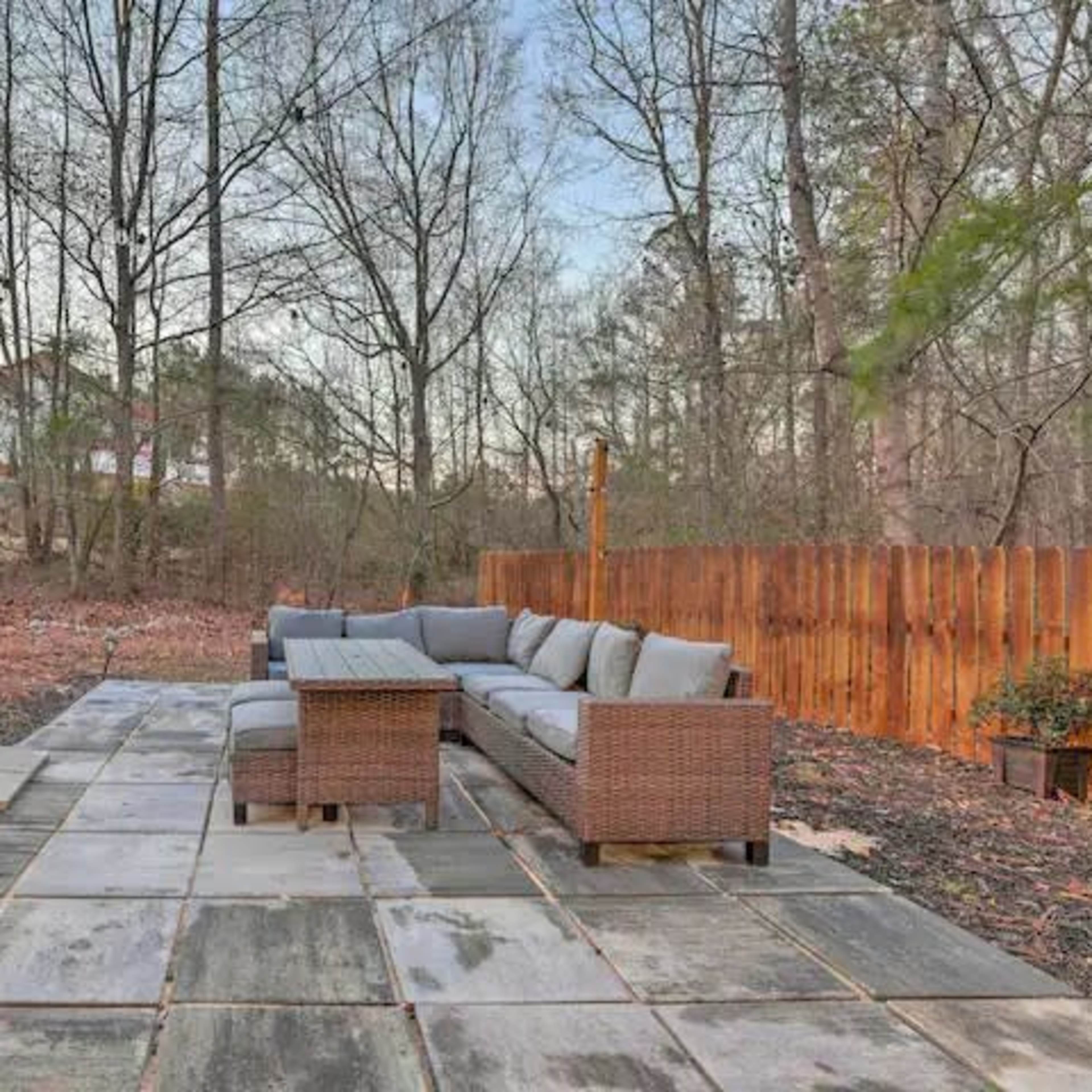 A patio area features a sectional sofa and a coffee table, surrounded by a landscaped yard with trees and a wooden fence in the background.
