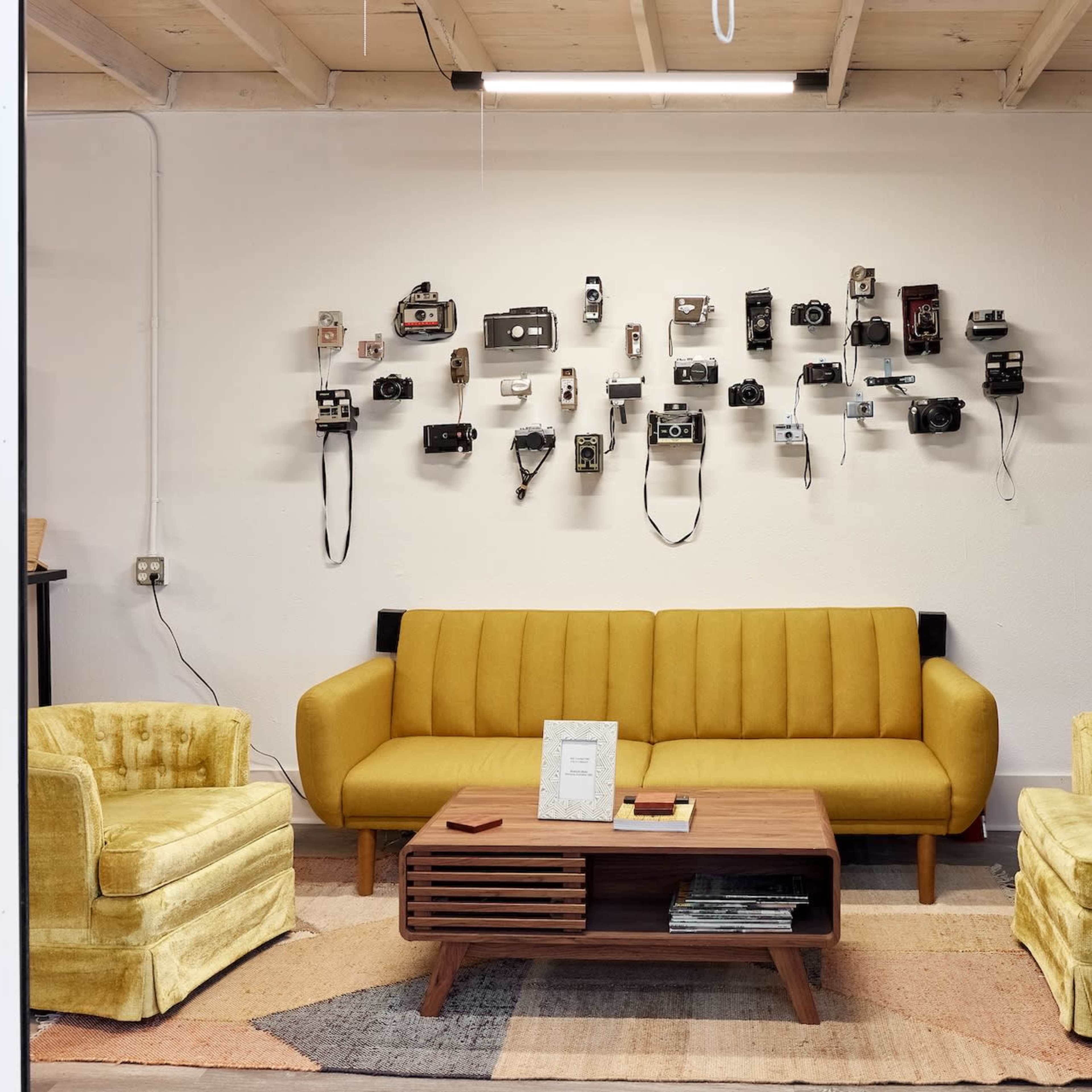 A cozy sitting area featuring a yellow sofa and two matching armchairs facing a wooden coffee table, with a wall adorned with various vintage cameras.