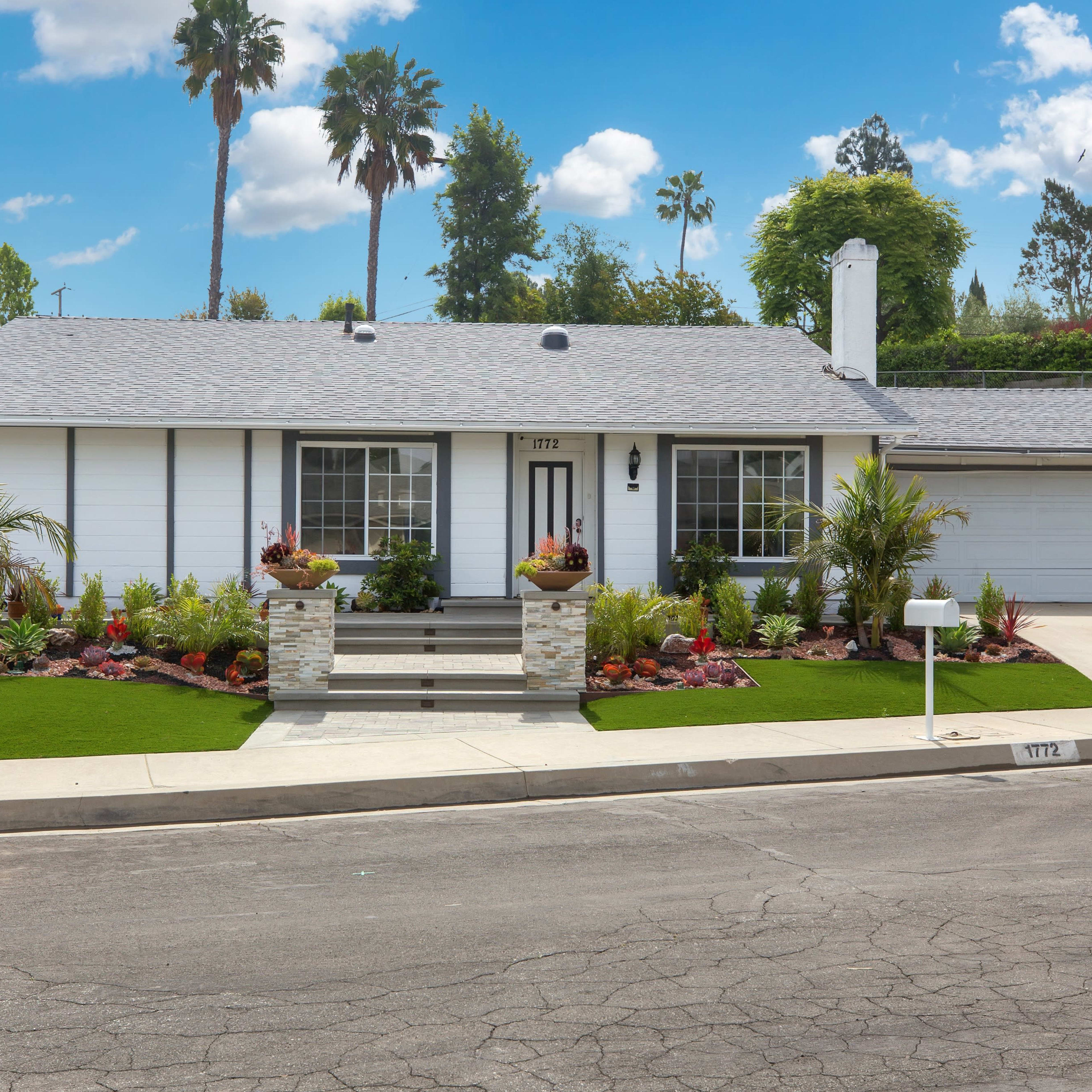 A single-story house with a landscaped front yard, featuring a stone pathway, flower beds, and a driveway.