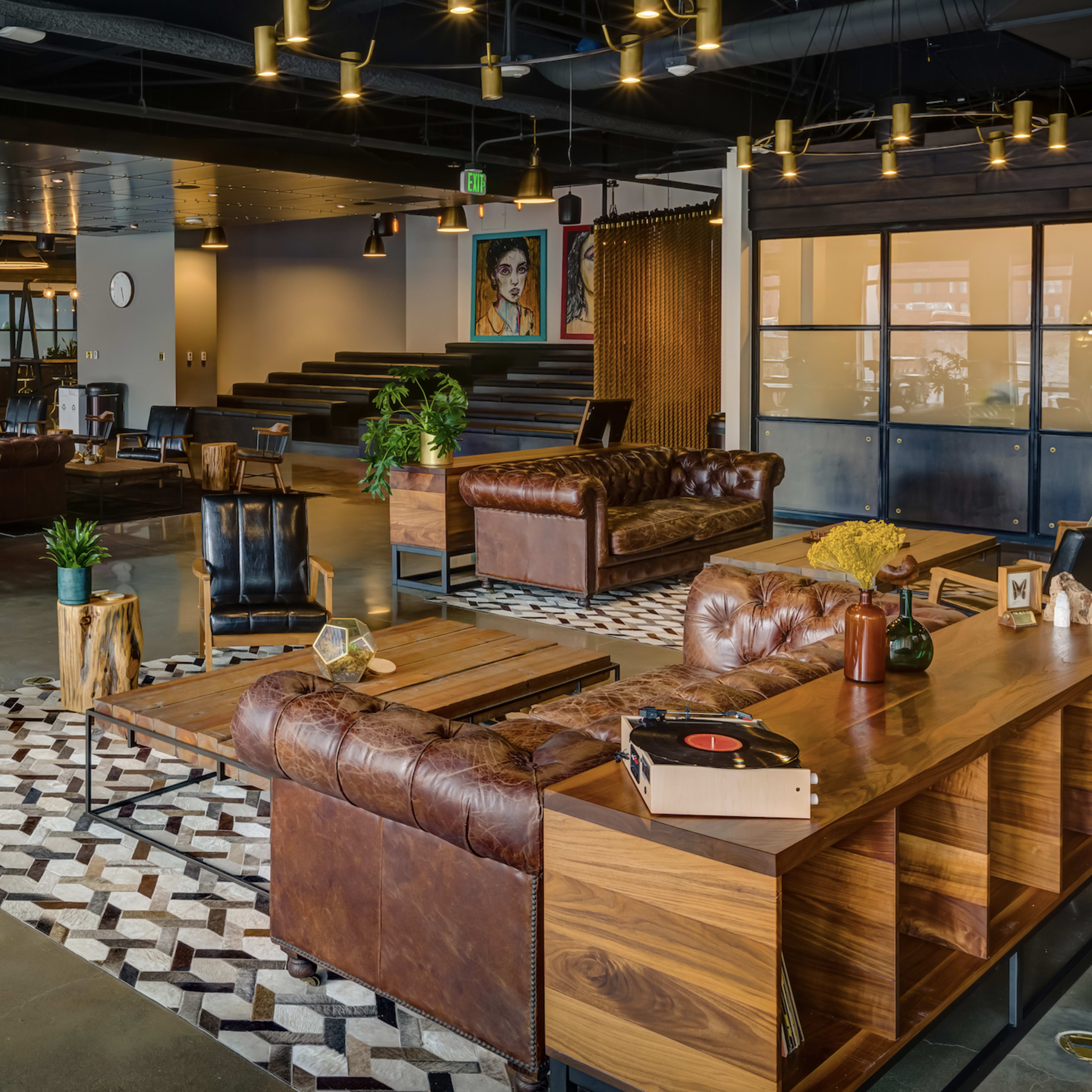 A spacious modern office interior with leather sofas, wooden furniture, and various seating arrangements scattered across a polished concrete floor.