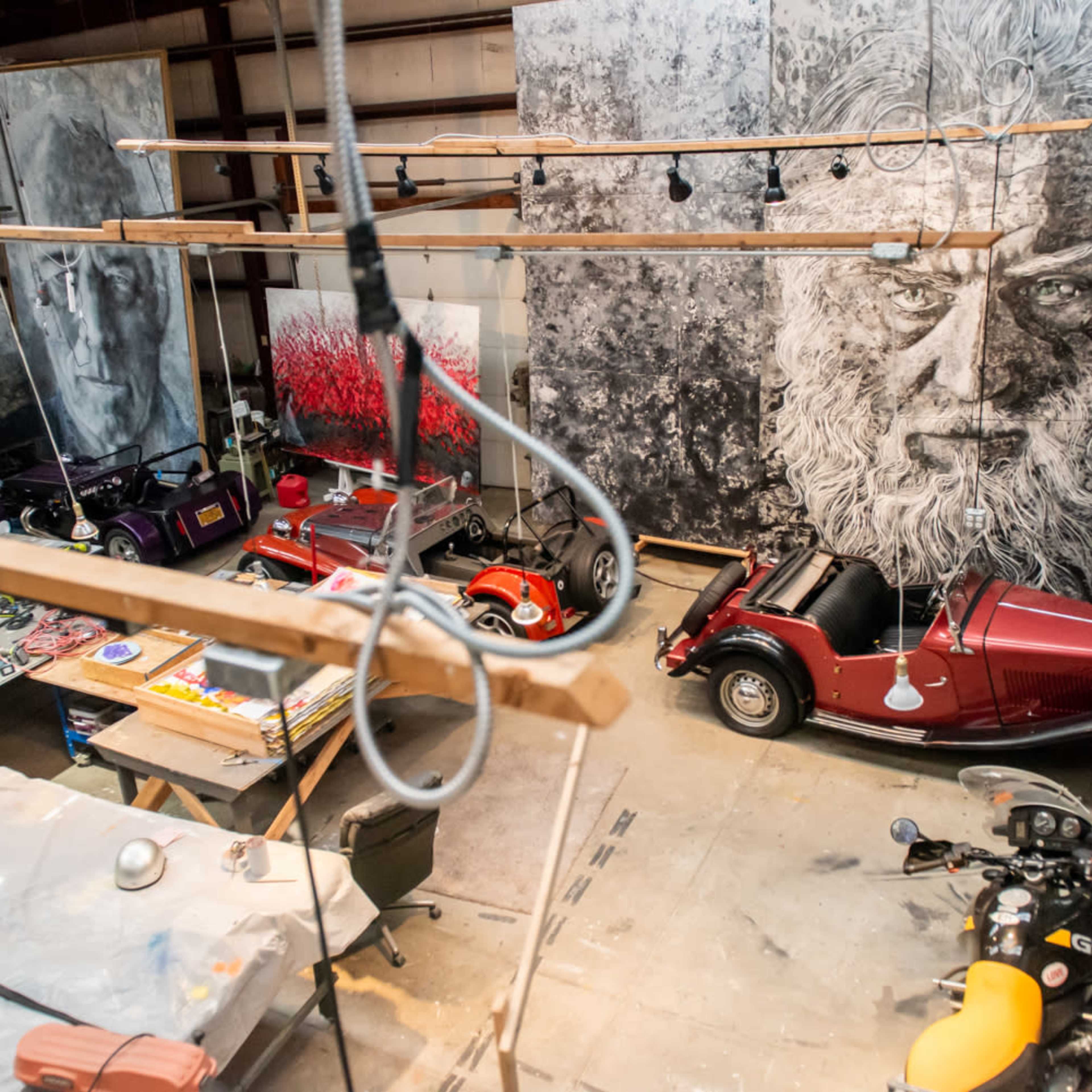 An artist's studio featuring multiple large paintings, several vintage cars, and a cluttered workspace.