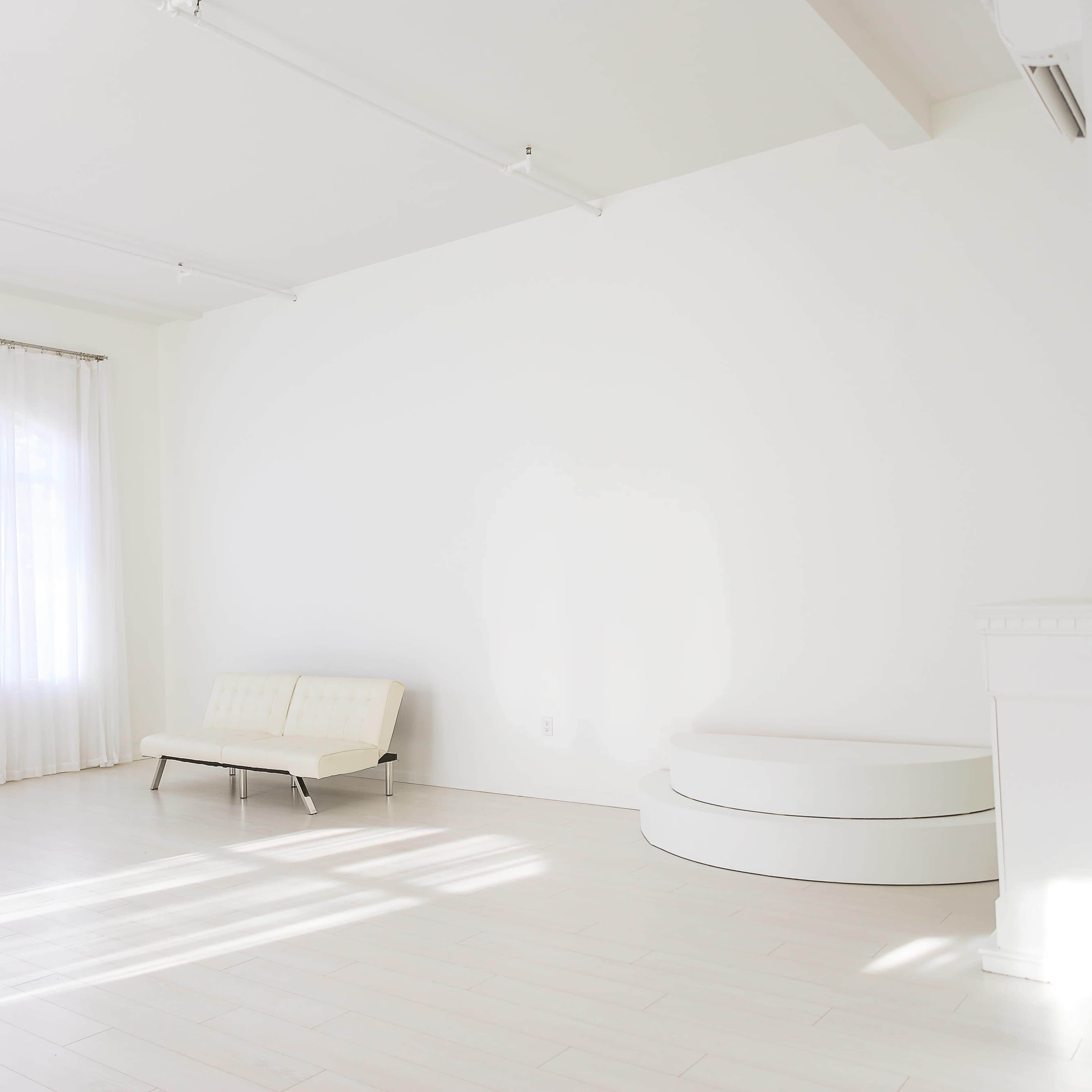 A bright, empty room features a white sofa, a curved white platform, and large windows covered with sheer curtains.
