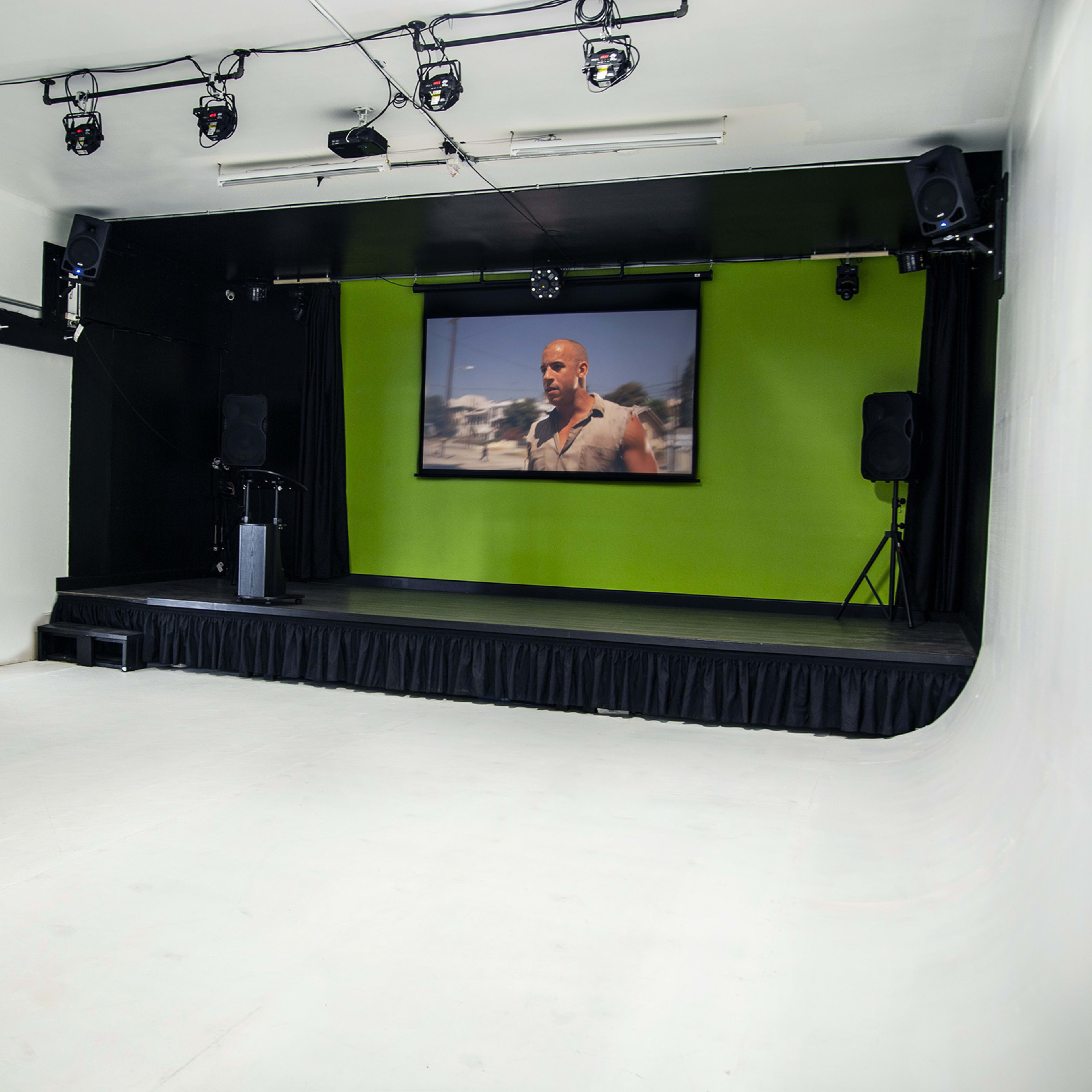 An empty stage with a green backdrop, a large screen displaying a scene, and audio equipment setup on either side.